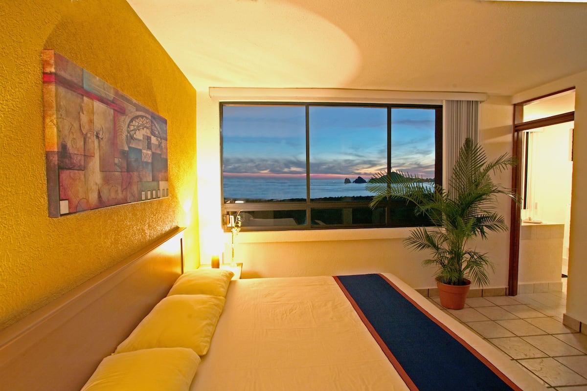 Habitación de hotel con un amplio ventanal que ofrece vistas al mar y al atardecer.