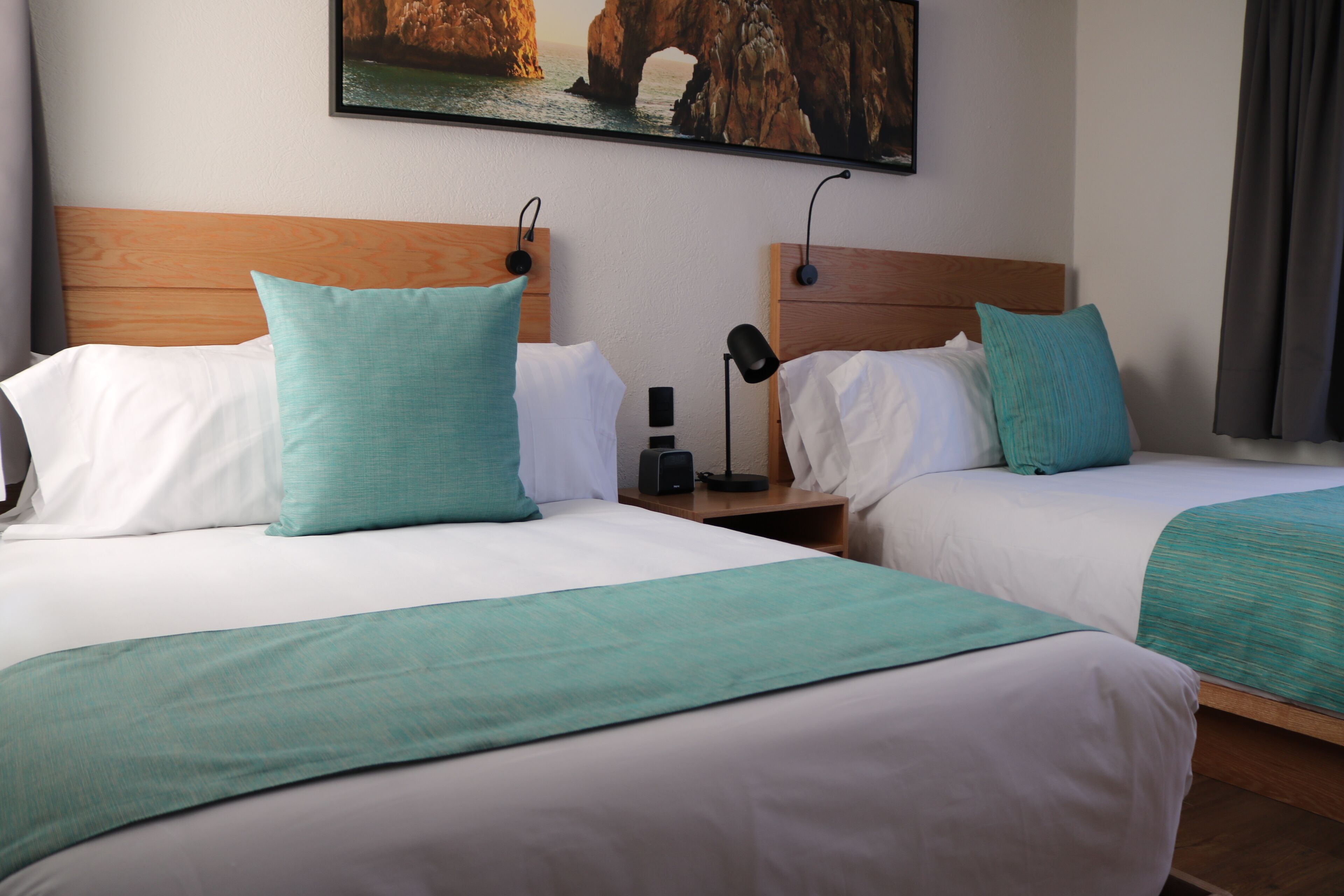 A hotel room with two beds, each with white bedding and teal pillows. There is a wooden headboard, a nightstand with a lamp, and a painting of a rocky landscape on the wall.