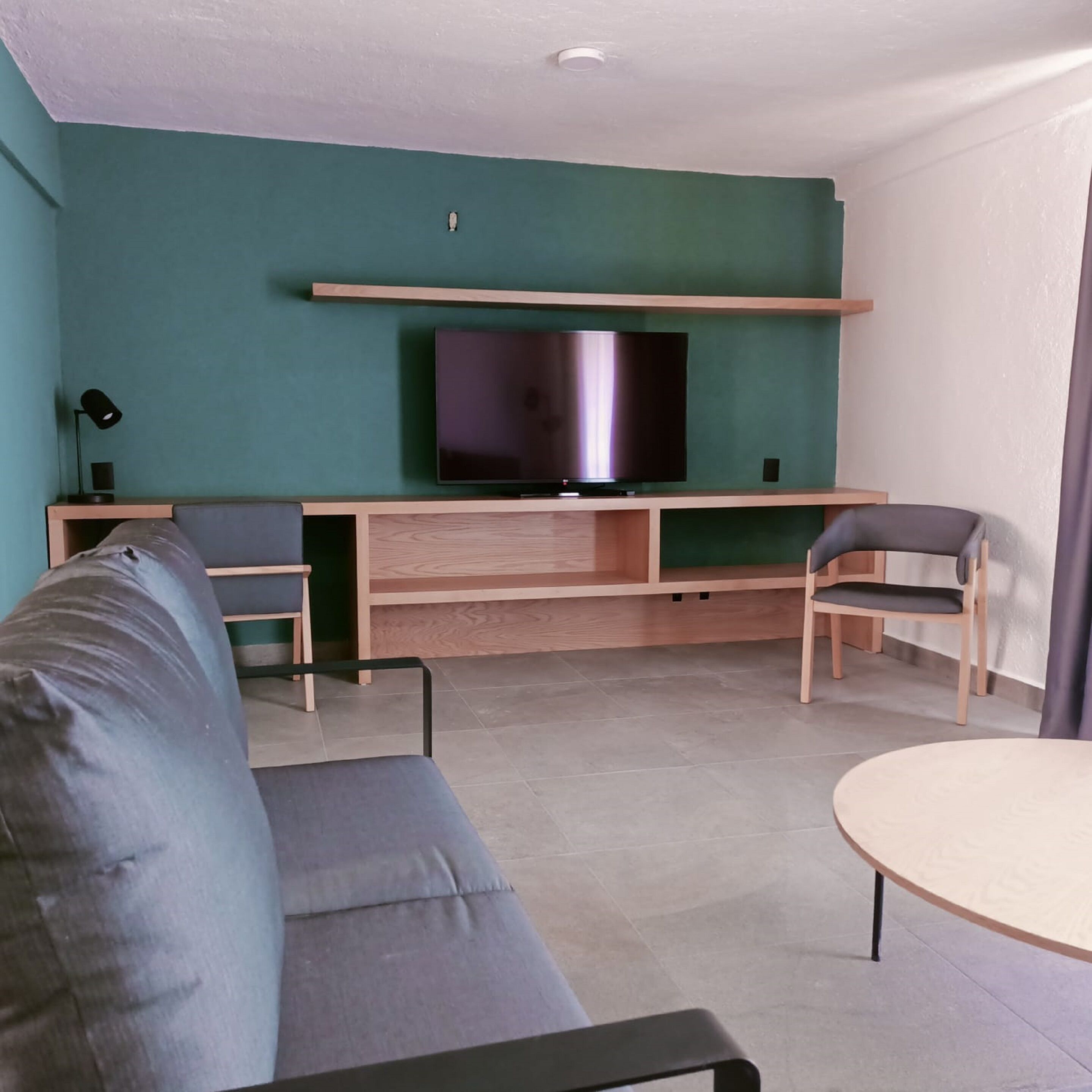 Una sala de estar moderna con una pared azul turquesa, un televisor de pantalla plana montado en un estante de madera, un sofá gris, una mesa de centro de madera y dos sillas de madera.