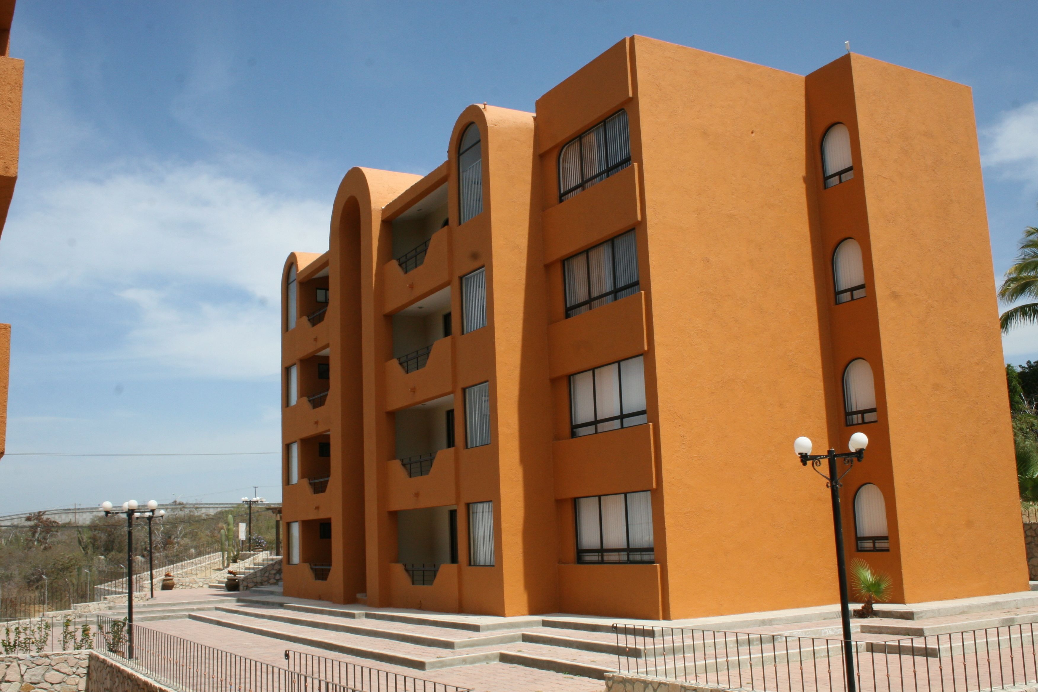 A multi-story building with balconies and large windows, featuring a flat roof and a paved area in front.