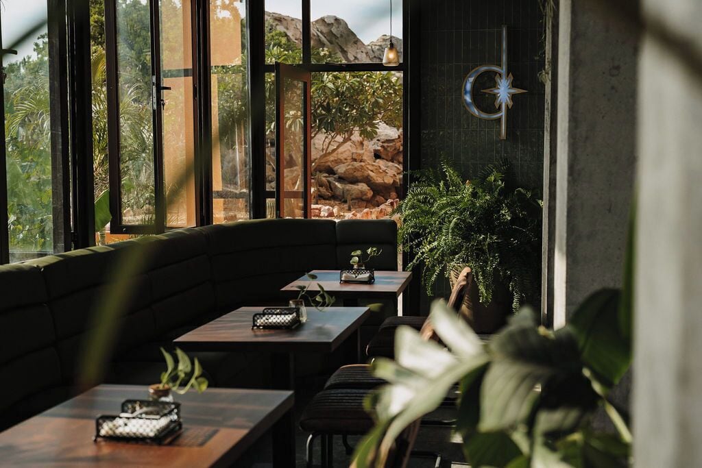 Een restaurant met een gebogen zithoek, grote ramen met uitzicht op rotsen en groen, en een decoratieve muur met een stervormig symbool.