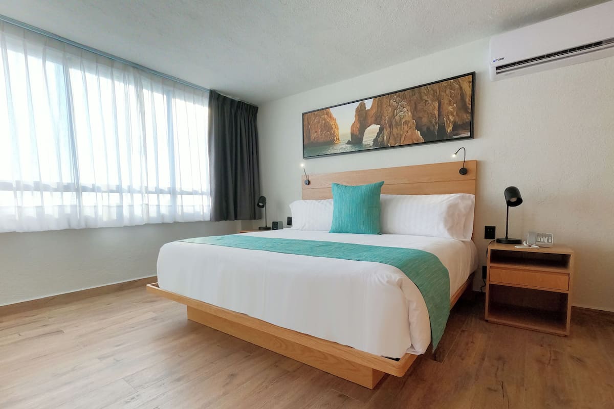 Habitación de hotel ordenada con una cama grande, una mesita de noche con lámpara, un cuadro en la pared y una ventana con cortinas.