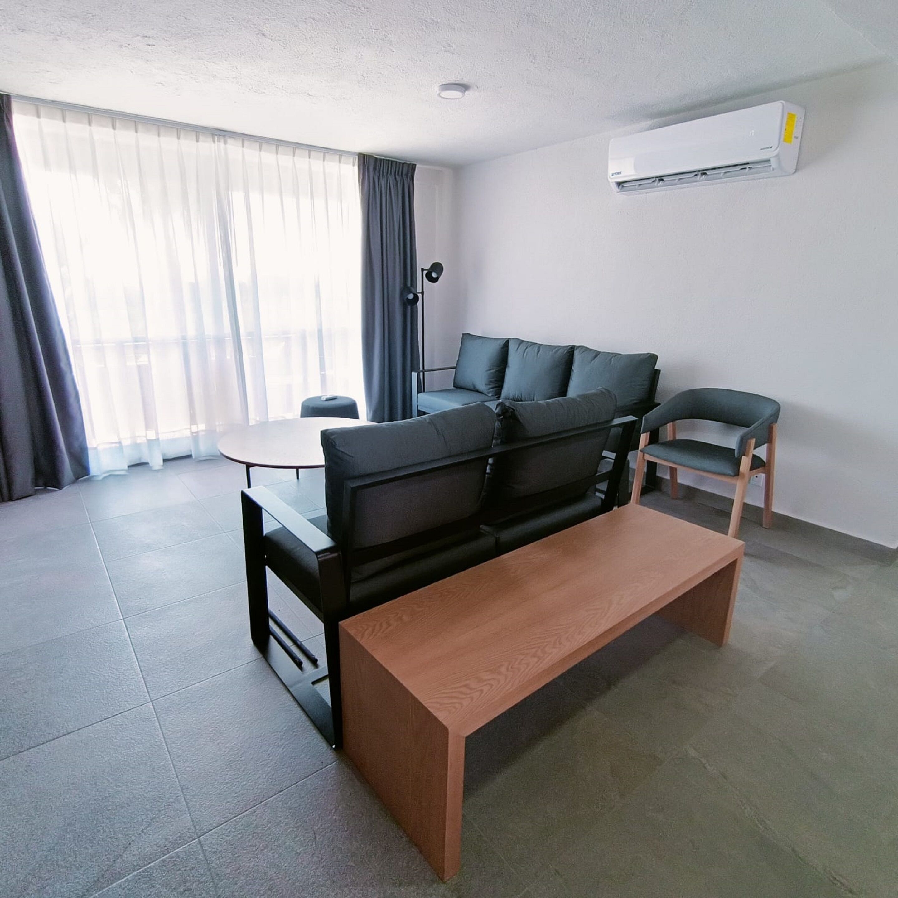 Un salon moderne avec un canapé noir, une table basse en bois, une petite table ronde et un fauteuil. Un appareil de climatisation est fixé au mur.