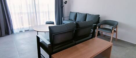 Una sala de estar moderna con un sofá negro, una mesa de centro de madera, una pequeña mesa redonda y una silla. Un equipo de aire acondicionado está montado en la pared.