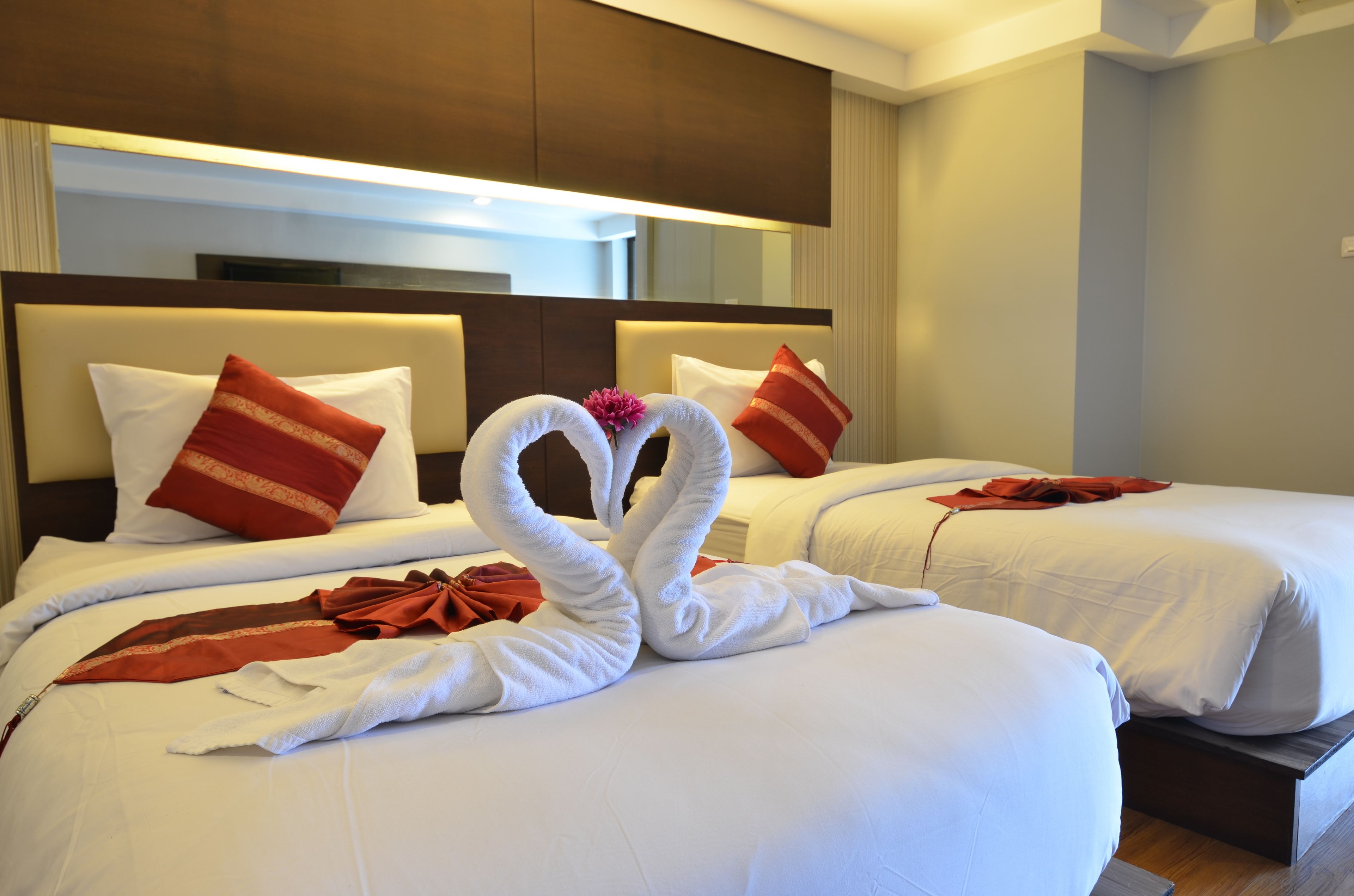 A hotel room with two beds, each with white linens and red pillows. There are two towels arranged in the shape of swans on the bed in the foreground.