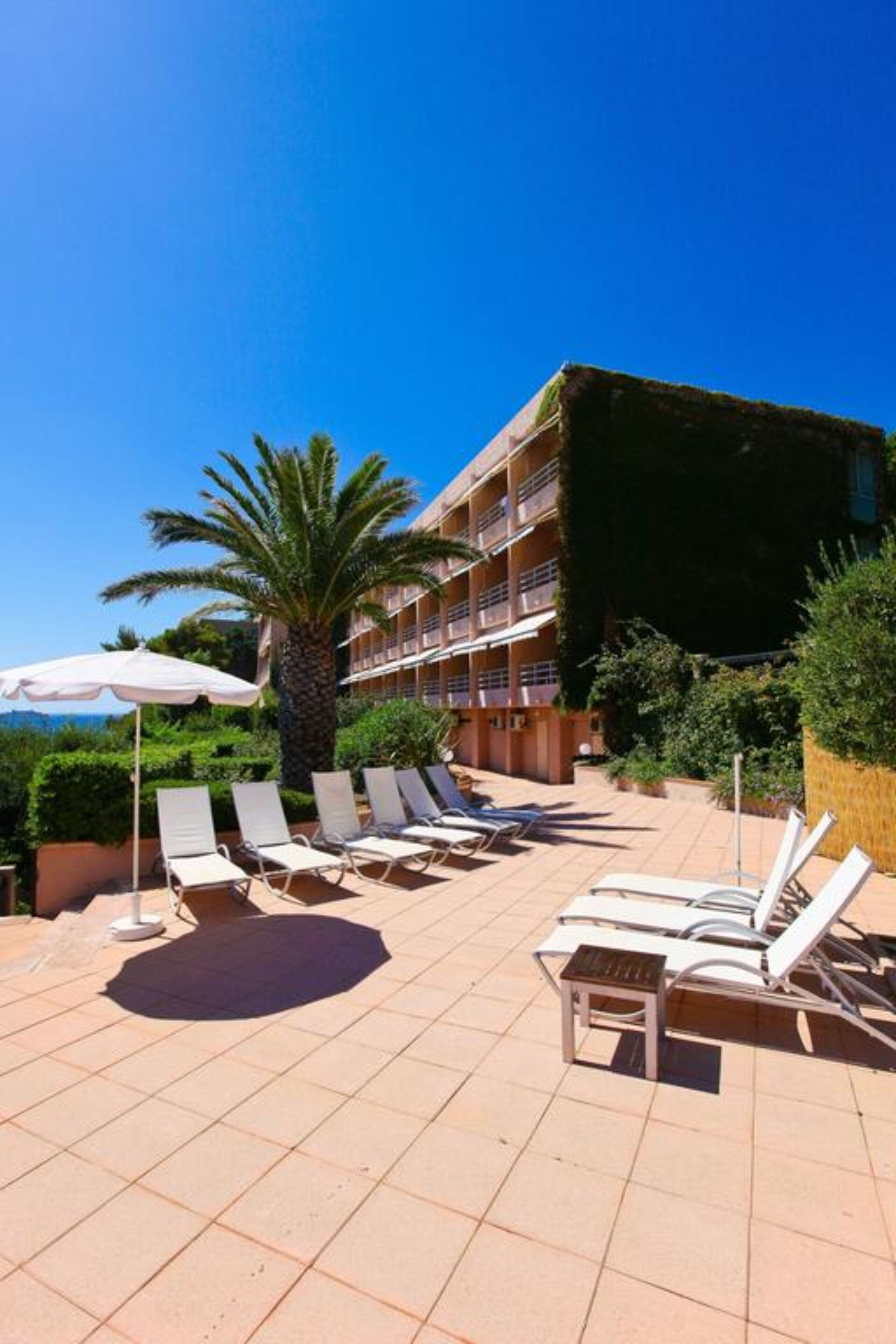 A hotel with a pool area featuring sun loungers and umbrellas, surrounded by greenery and a clear blue sky.