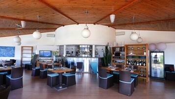 A spacious indoor area with wooden ceiling beams, multiple tables and chairs, a bar area with shelves of bottles, and a large window providing natural light.
