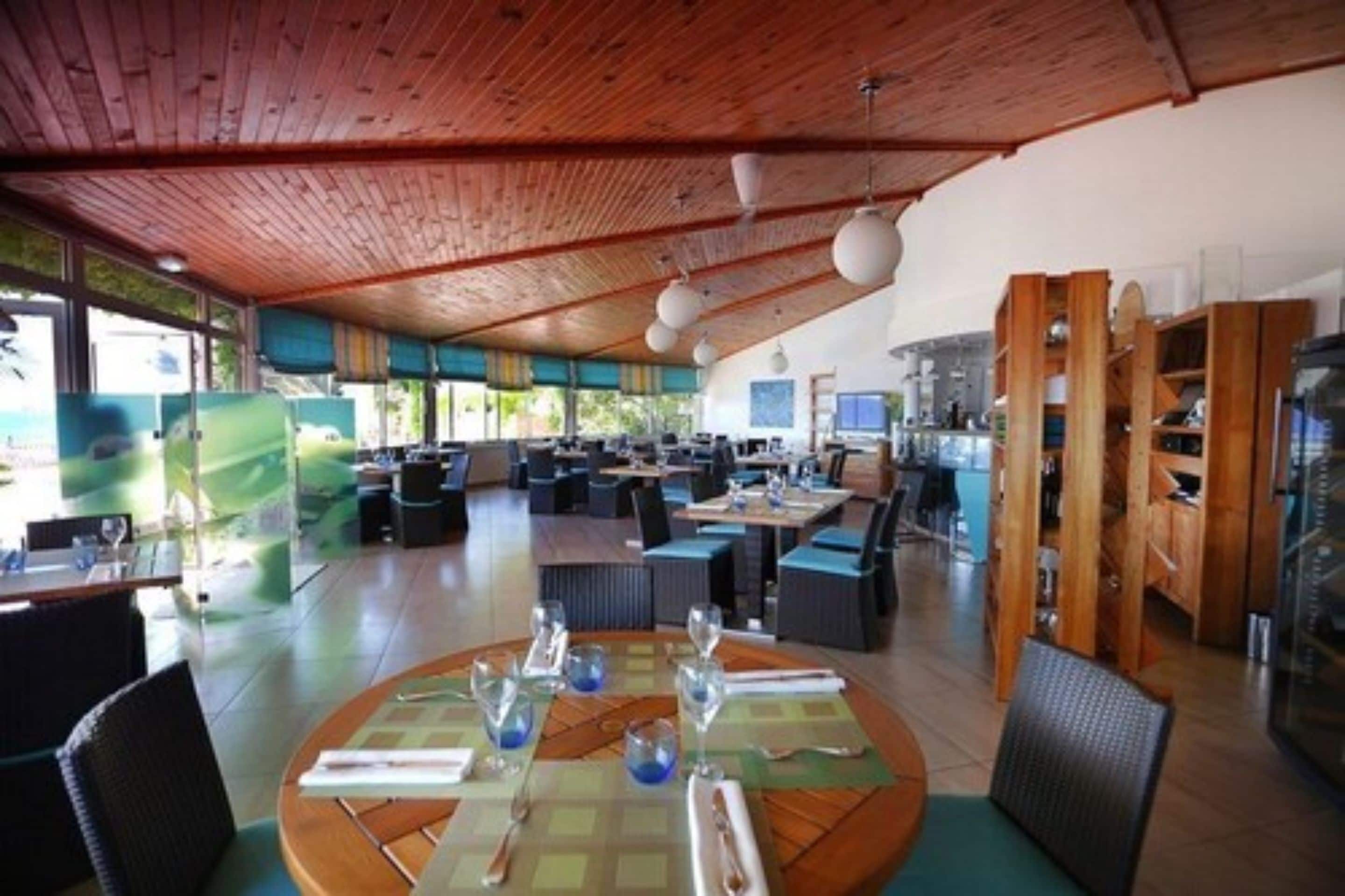 Un restaurante espacioso con vigas de madera en el techo, mesas redondas con manteles individuales azules y vistas al exterior.