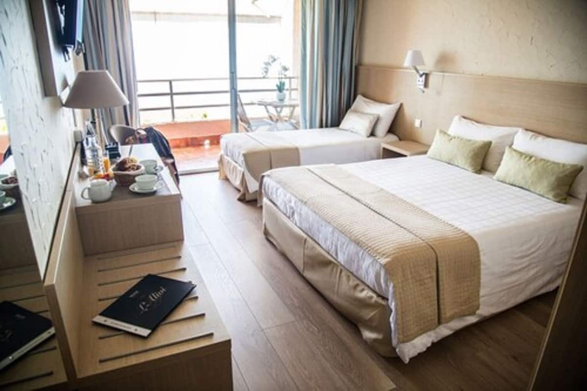 A hotel room with two beds, a desk with a lamp, and a view of the outdoors.
