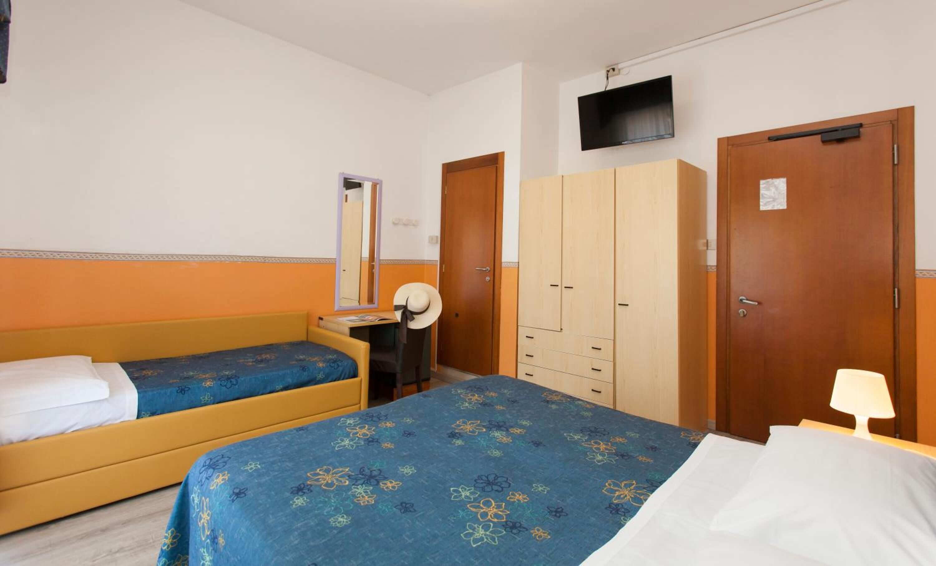 Ett hotellrum med två separata sängar, en garderob i trä, ett skrivbord med en stol, en spegel och en tv som är monterad på väggen.