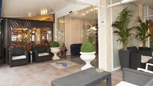 Un hall con muebles de mimbre, plantas en macetas y una entrada de vidrio que da a un restaurante.