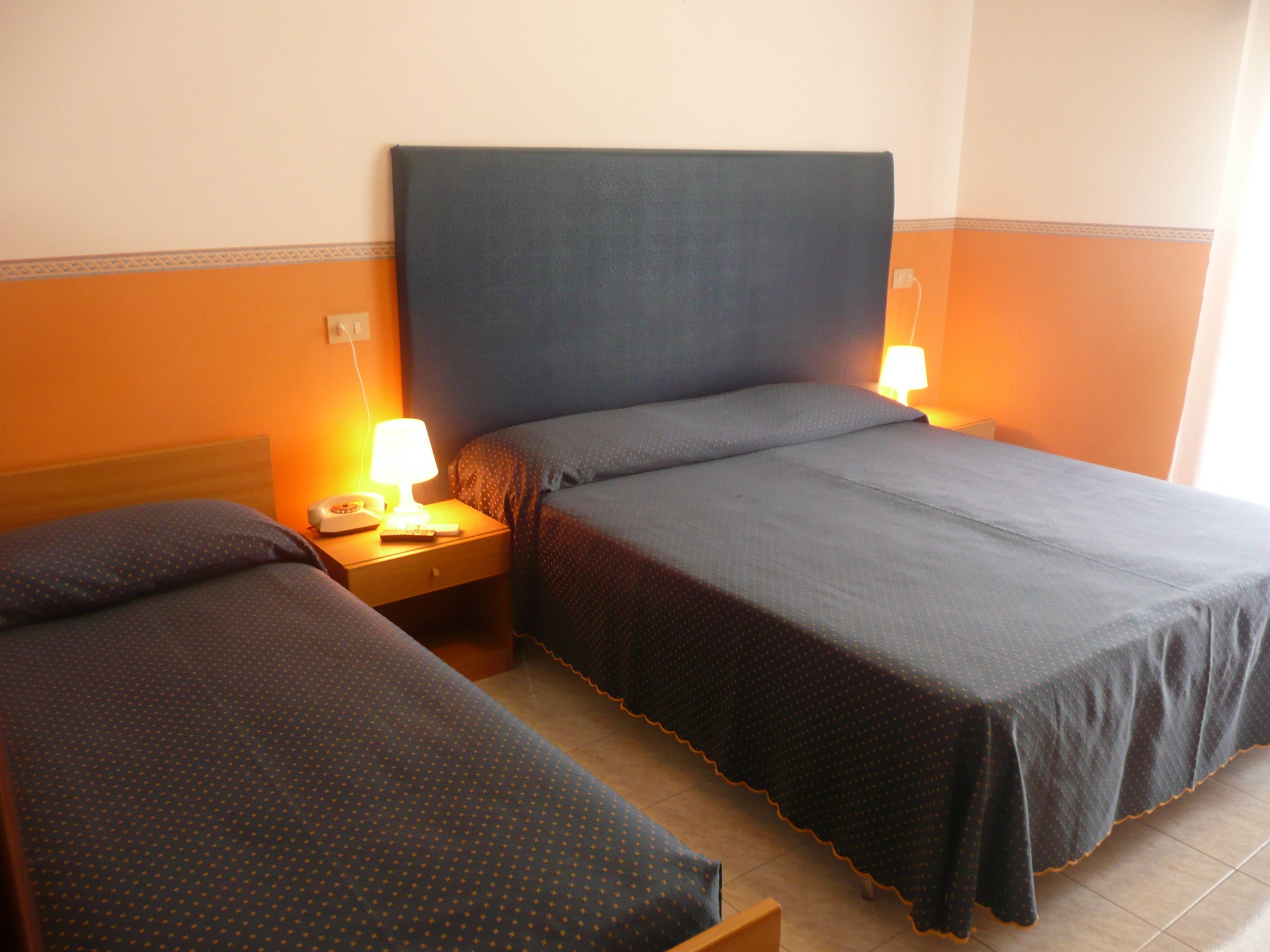 Quarto de hotel com duas camas de solteiro, mesa de cabeceira com abajur e telefone, e uma tomada na parede.