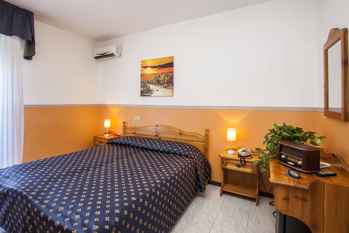 A hotel room with a bed, bedside lamps, a nightstand with a phone and radio, a wall-mounted air conditioner, and a painting of a sunset over a cityscape.