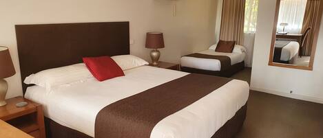 A hotel room with a large bed, a smaller bed, a desk, a chair, a lamp, a mirror, and an air conditioning unit.
