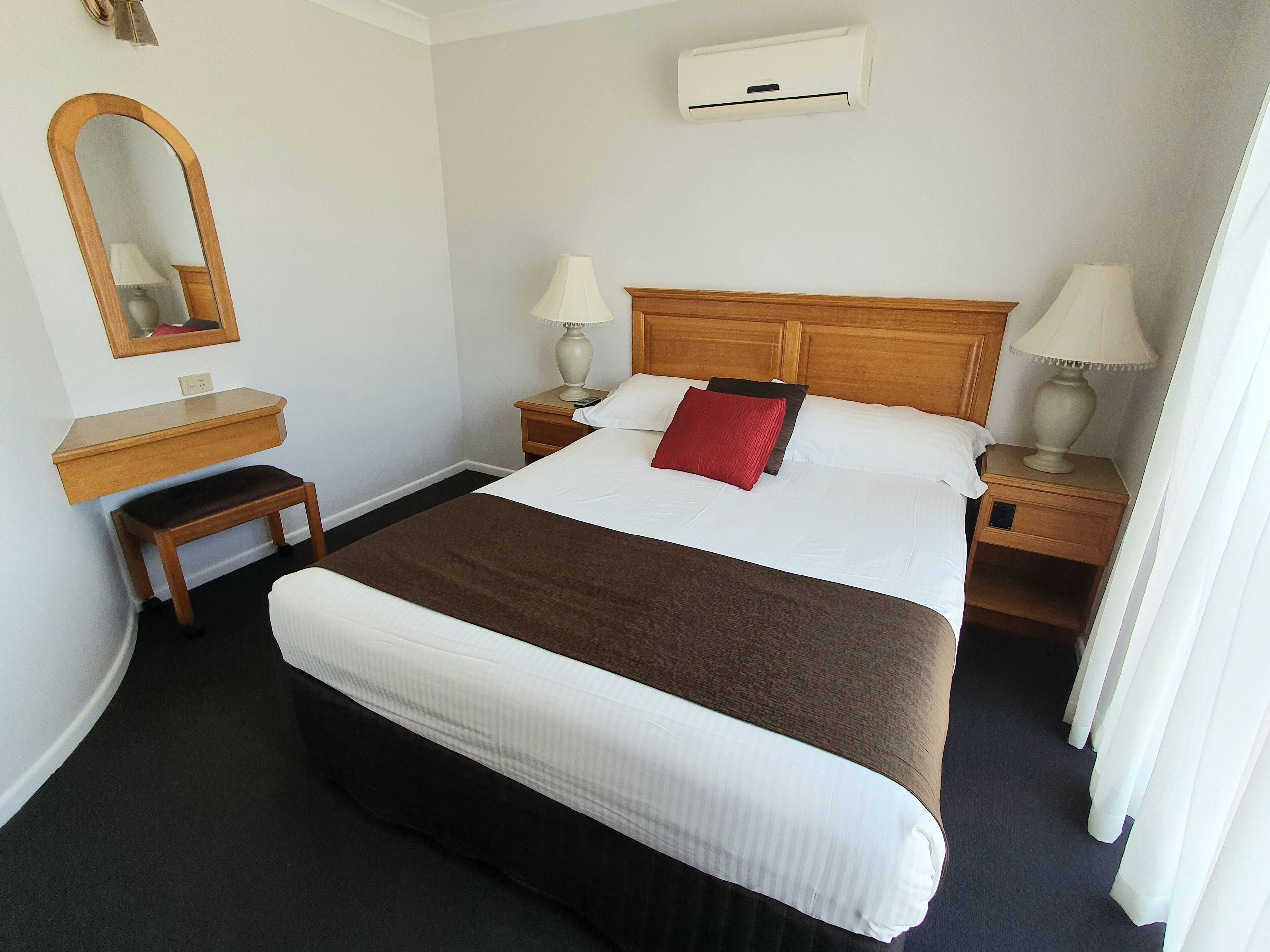 Une chambre d’hôtel avec un grand lit, deux tables de chevet avec des lampes, une tête de lit en bois, un petit banc en bois, un miroir et un appareil de climatisation fixé au mur.