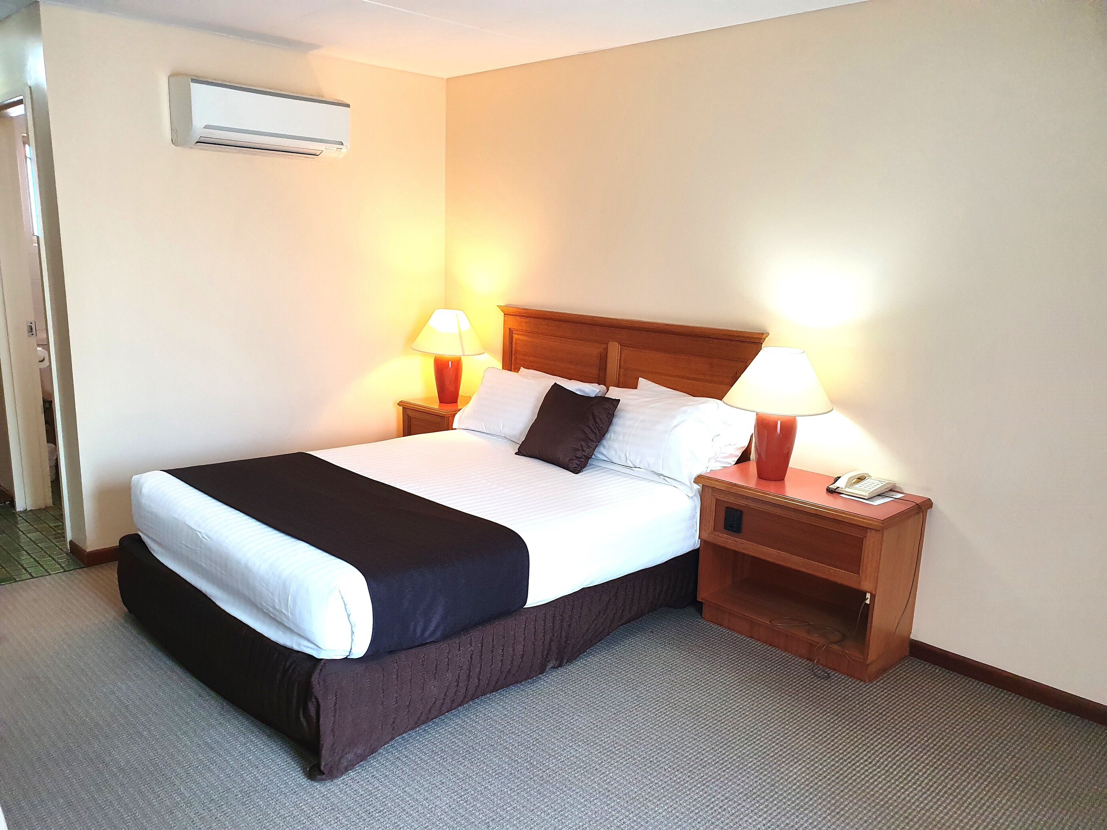 A hotel room with a bed, two bedside lamps, a nightstand, and an air conditioning unit.
