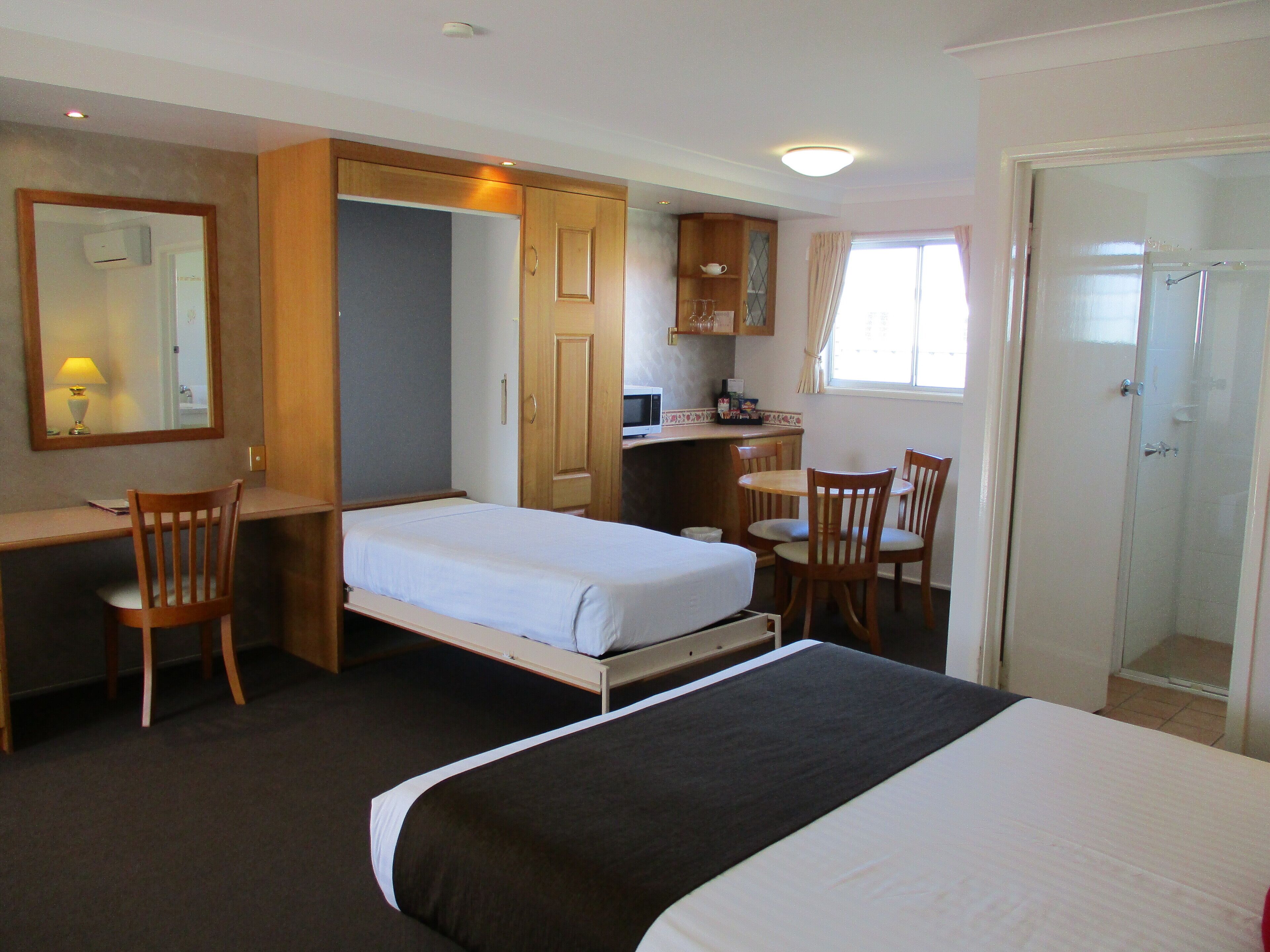 Quarto de hotel compacto com cama, escrivaninha, cadeira, pequeno espaço de cozinha e área de chuveiro.