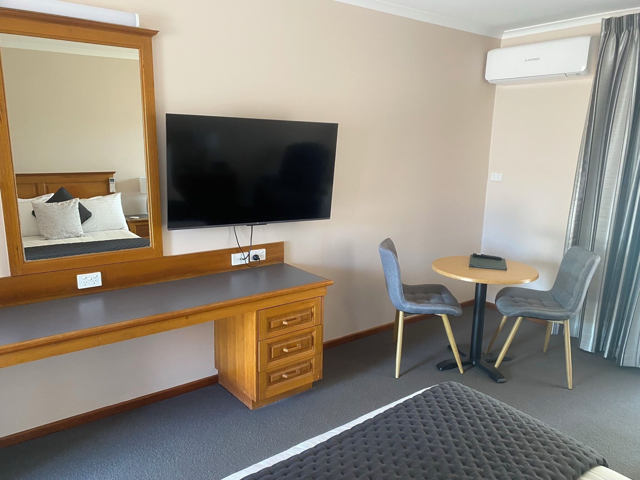 A hotel room with a wooden desk, a flat-screen TV, two chairs, a small round table, and an air conditioning unit.
