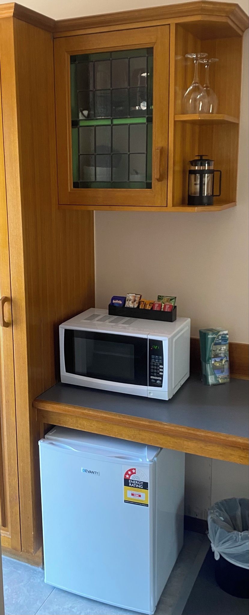 Una kitchenette con microondas, un refrigerador pequeño y un armario con puertas de vidrio que contiene utensilios de cocina.