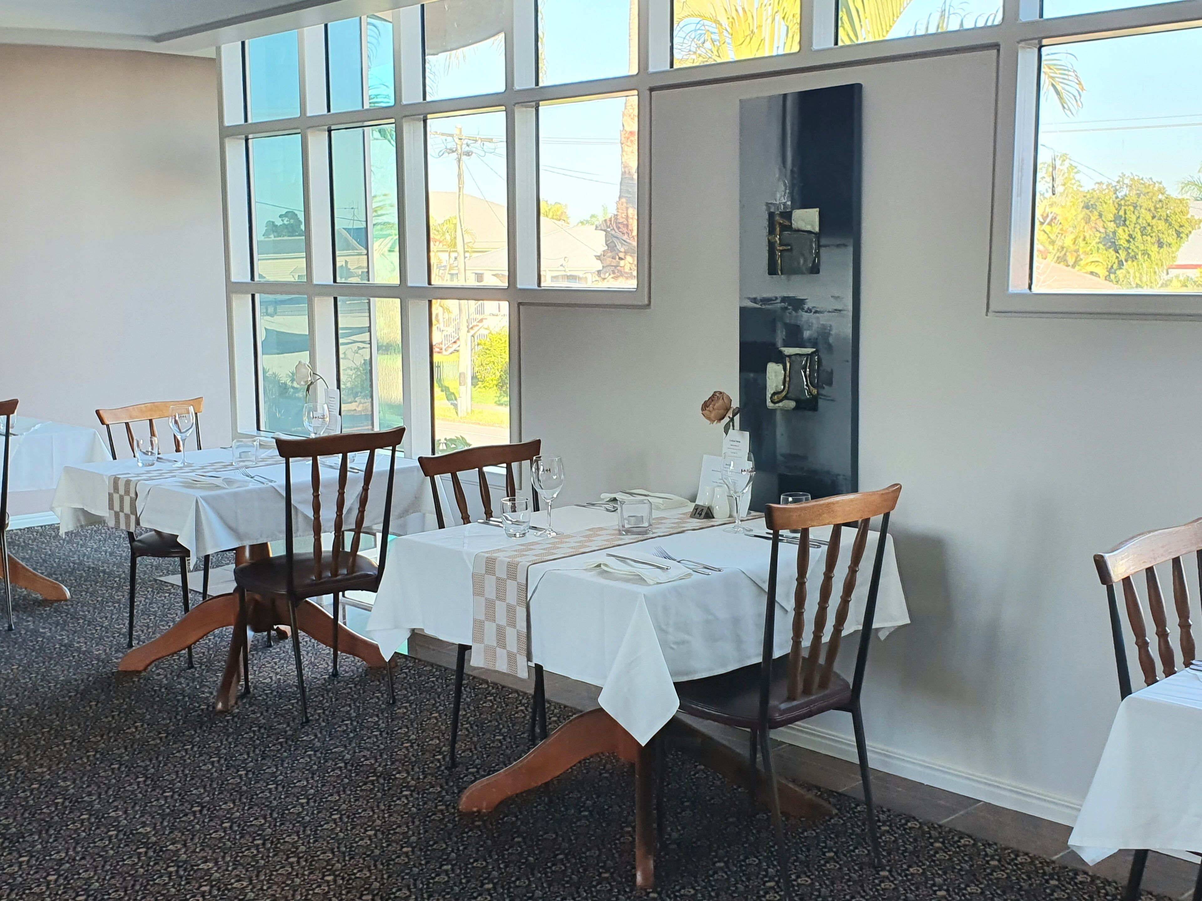 A dining area with tables set for four, each with a white tablecloth, wooden chairs, and a vase with a single flower.