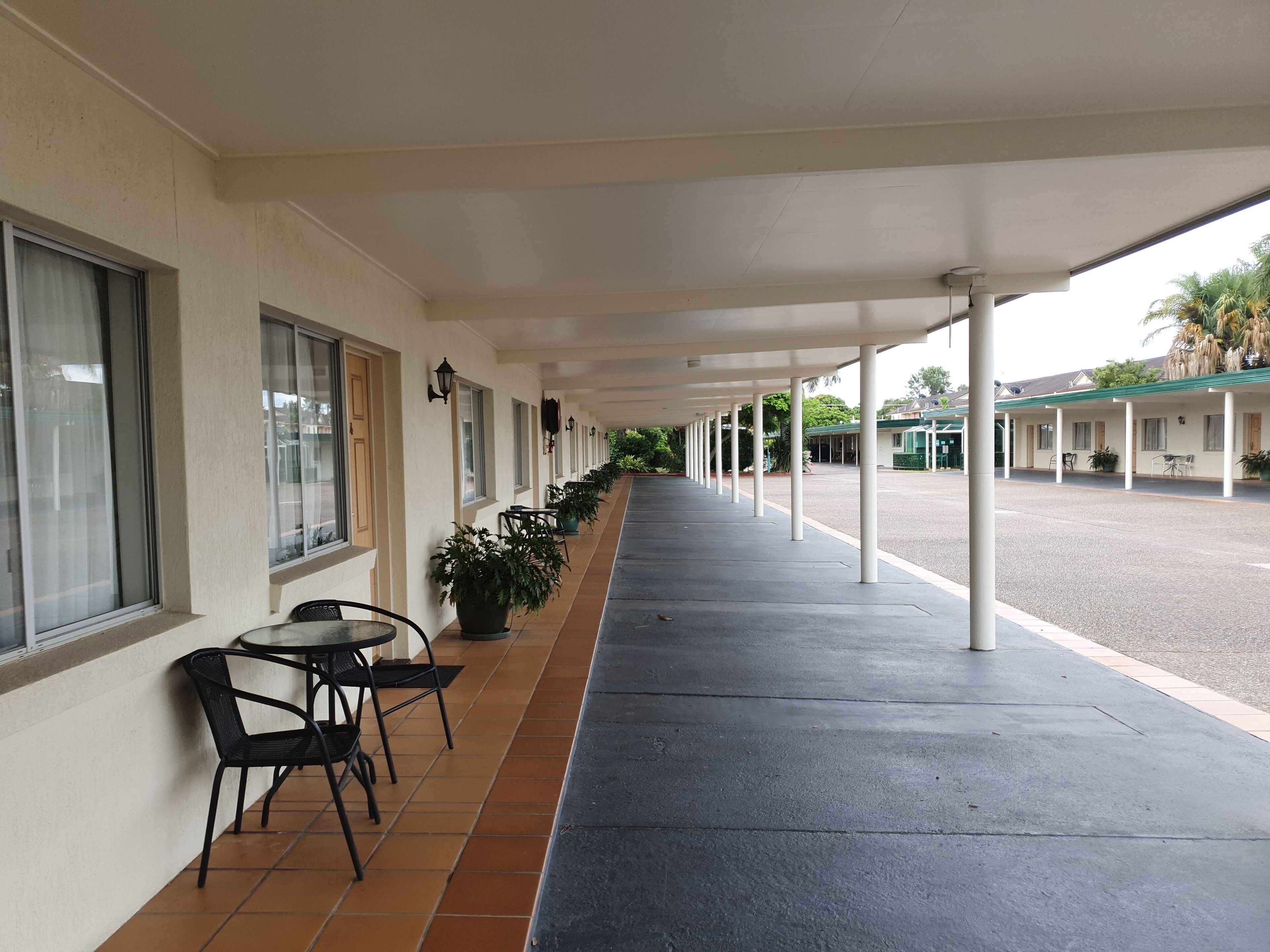 A covered walkway with a row of windows and chairs, leading to a building with a green roof.