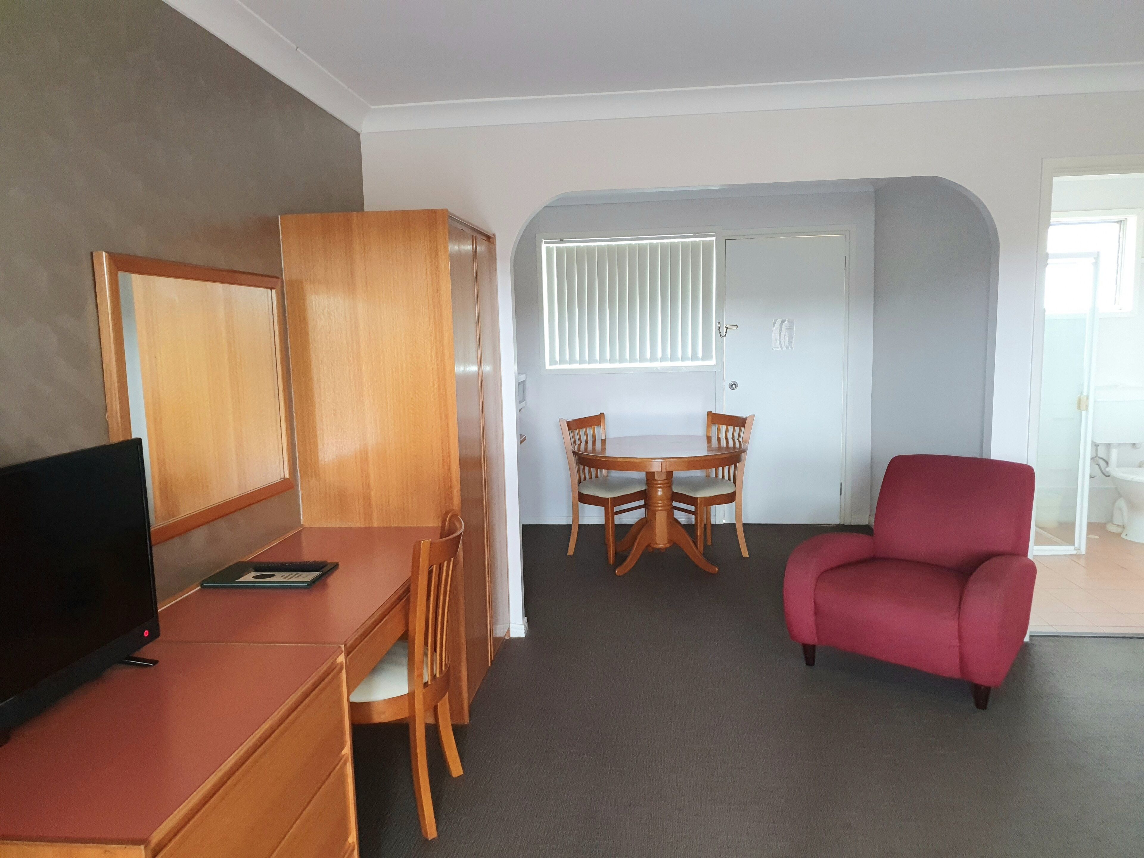 Habitación de hotel con un escritorio de madera, un televisor, un sillón rojo, una mesa de comedor con sillas y vistas al baño a través de una puerta abierta.
