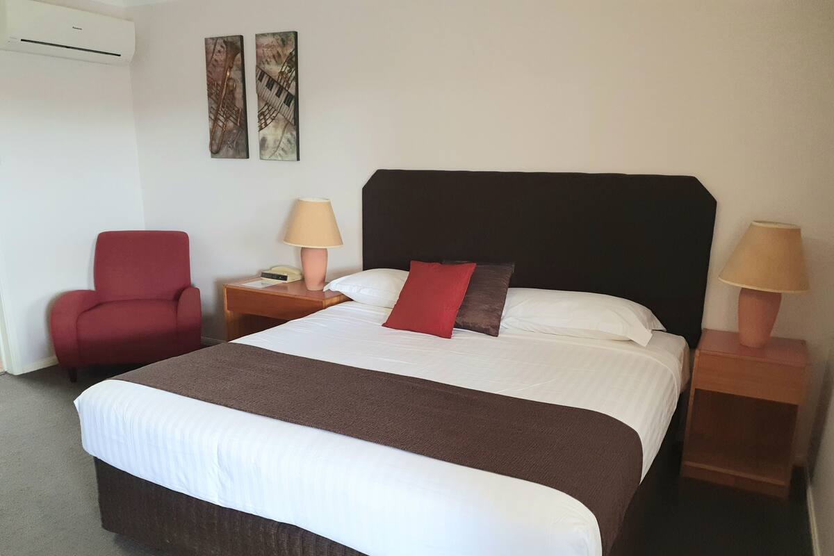 A hotel room with a large bed, two bedside lamps, a red armchair, a small table with a lamp, and two wall art pieces.