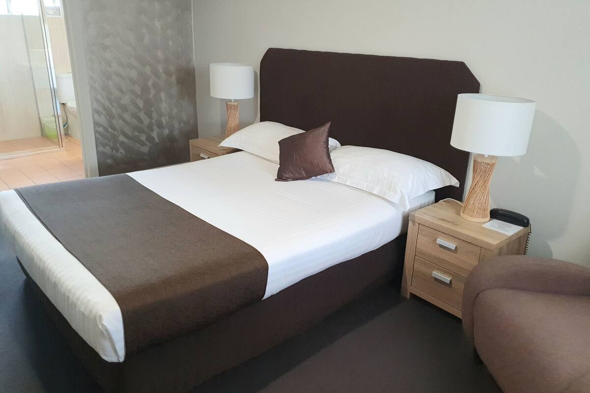 A neatly made bed with a brown headboard, two bedside tables with lamps, a wooden nightstand, and a brown chair.
