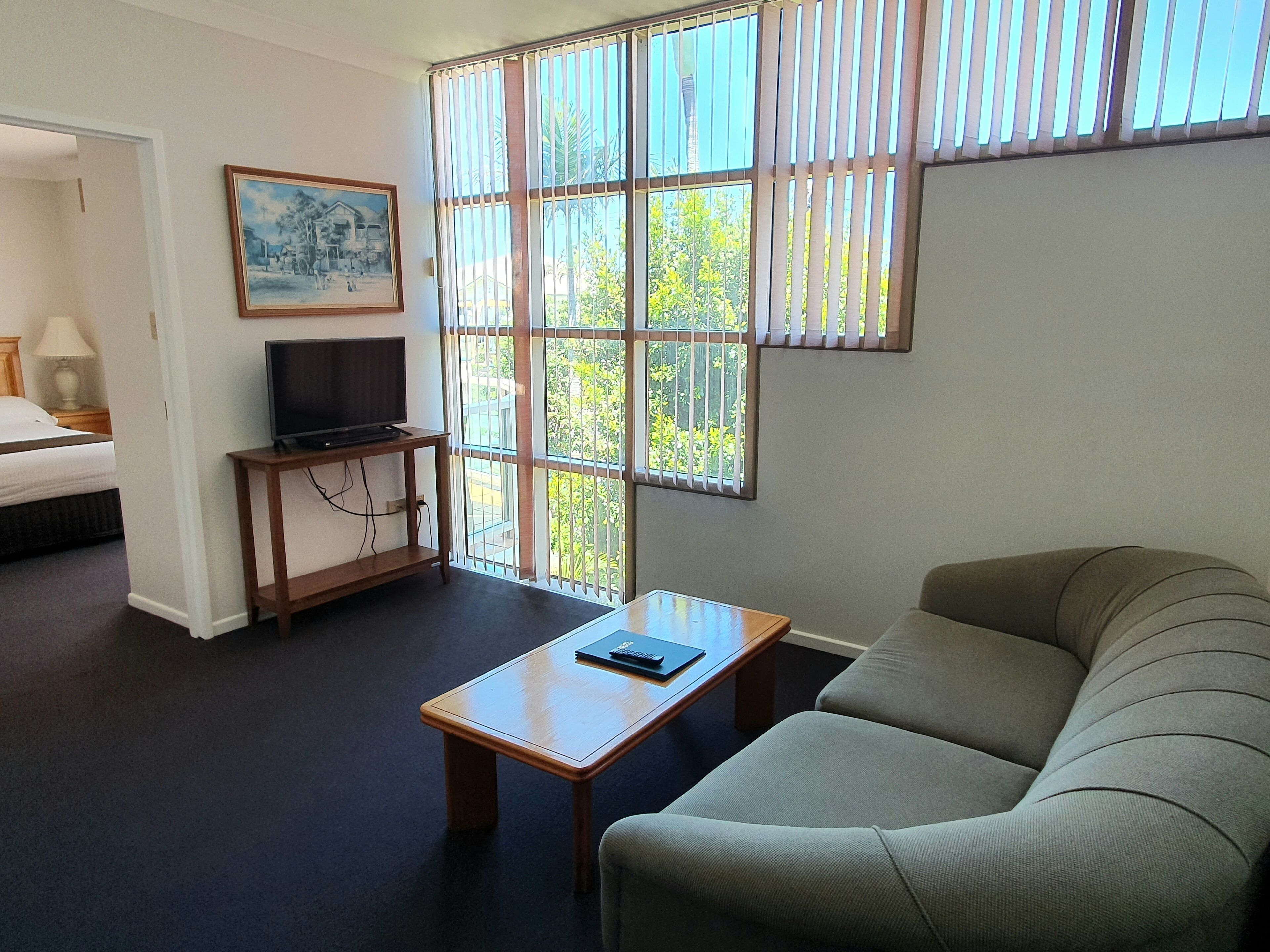 Habitación de hotel con cama, televisor sobre un mueble de madera, sofá, mesa de centro y ventana con persianas.