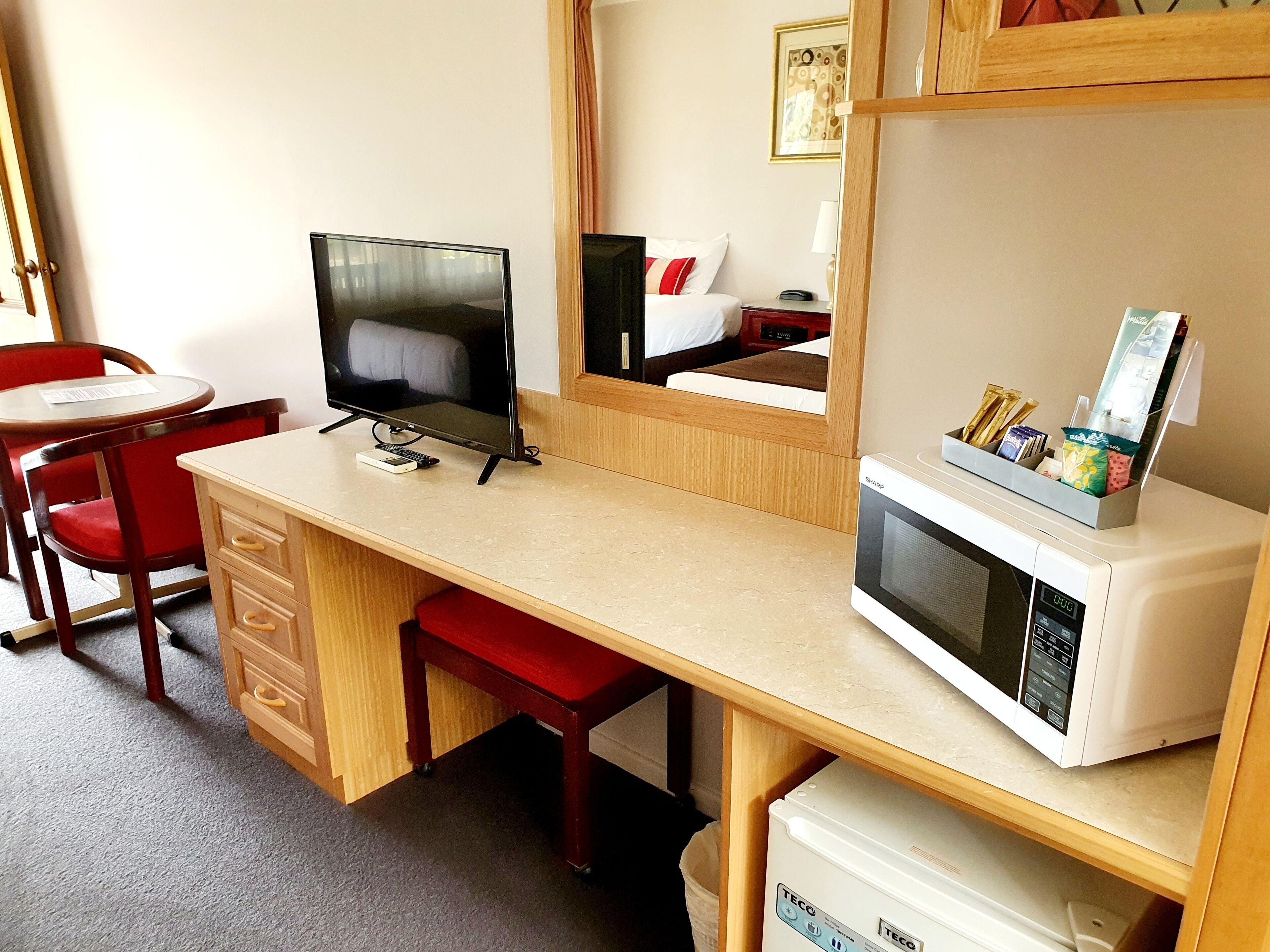 Quarto de hotel compacto com uma escrivaninha de madeira, televisão, micro-ondas e uma pequena área de cozinha.