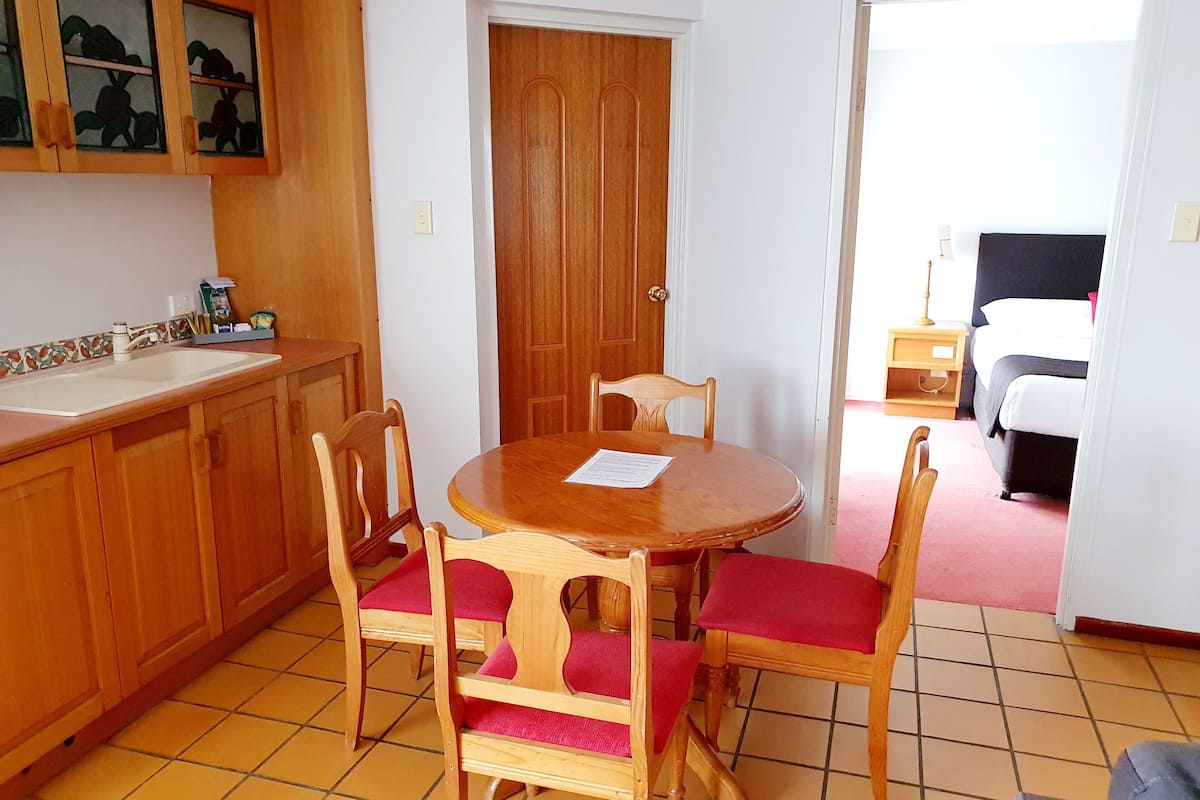 Une petite pièce bien éclairée, avec une table et des chaises en bois, un coin cuisine comprenant un évier et des placards, et une porte donnant sur une chambre à coucher avec un lit et une table de chevet.