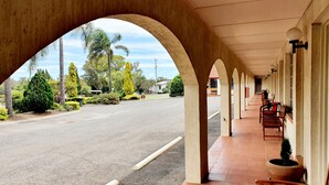 A covered walkway with a view of a road and greenery, featuring a row of benches and potted plants. - McNevin's Warwick Motel (Warwick)