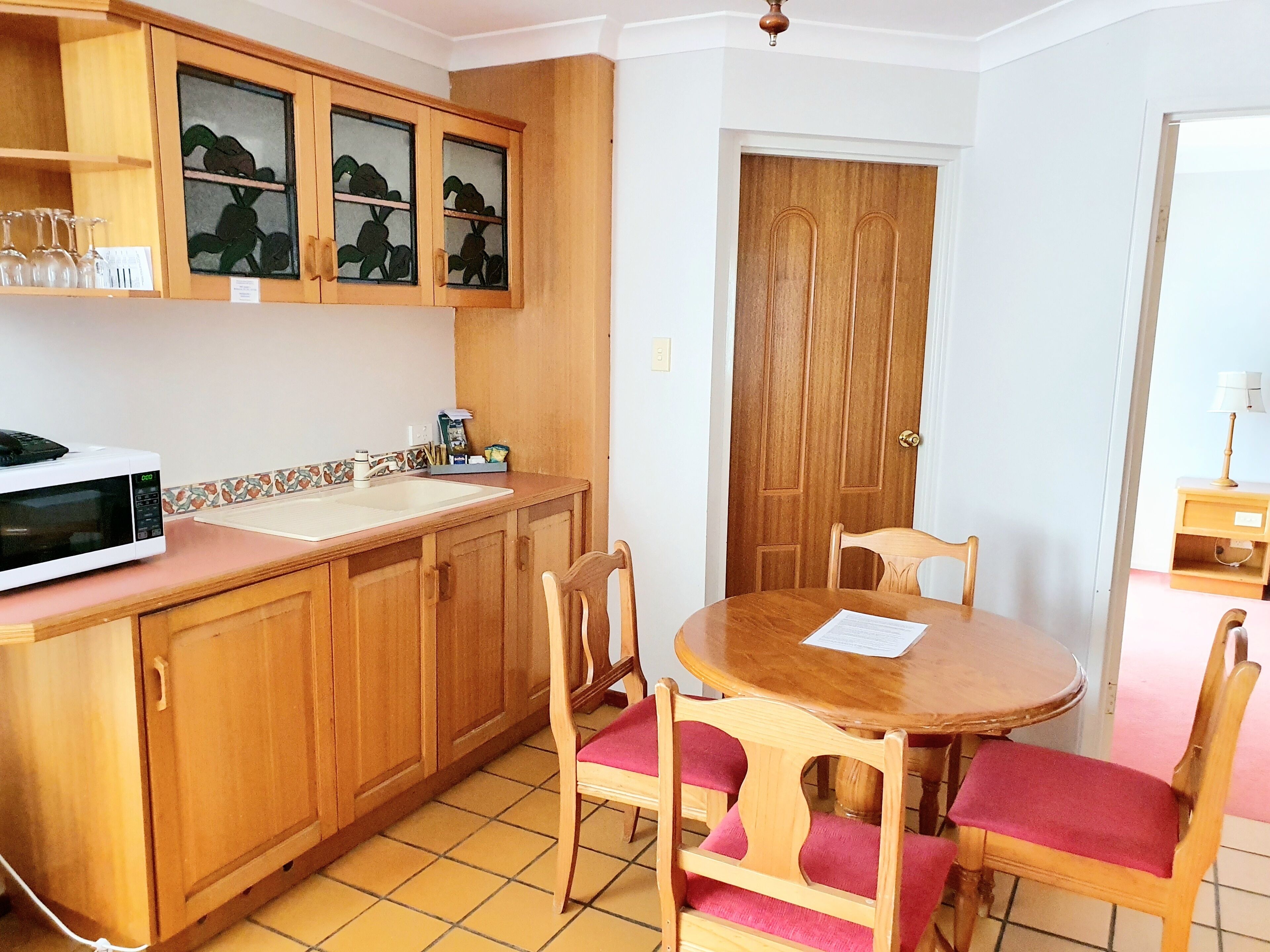 Uma cozinha compacta com armários de madeira, micro-ondas e pia. Há uma mesa de jantar com cadeiras e uma porta ao fundo.