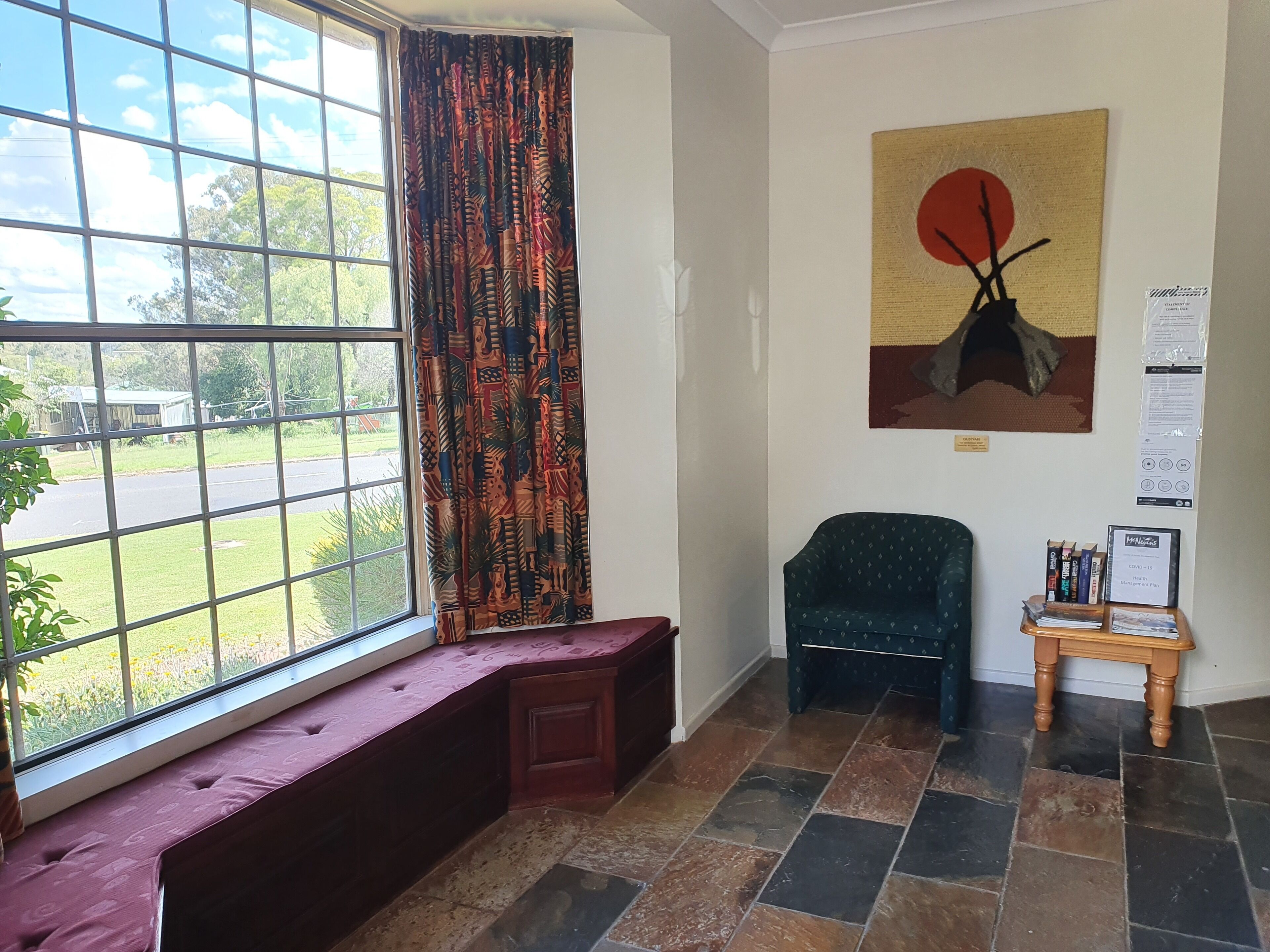 A room with a bench by a window, a chair, a small table with books, and a painting on the wall.