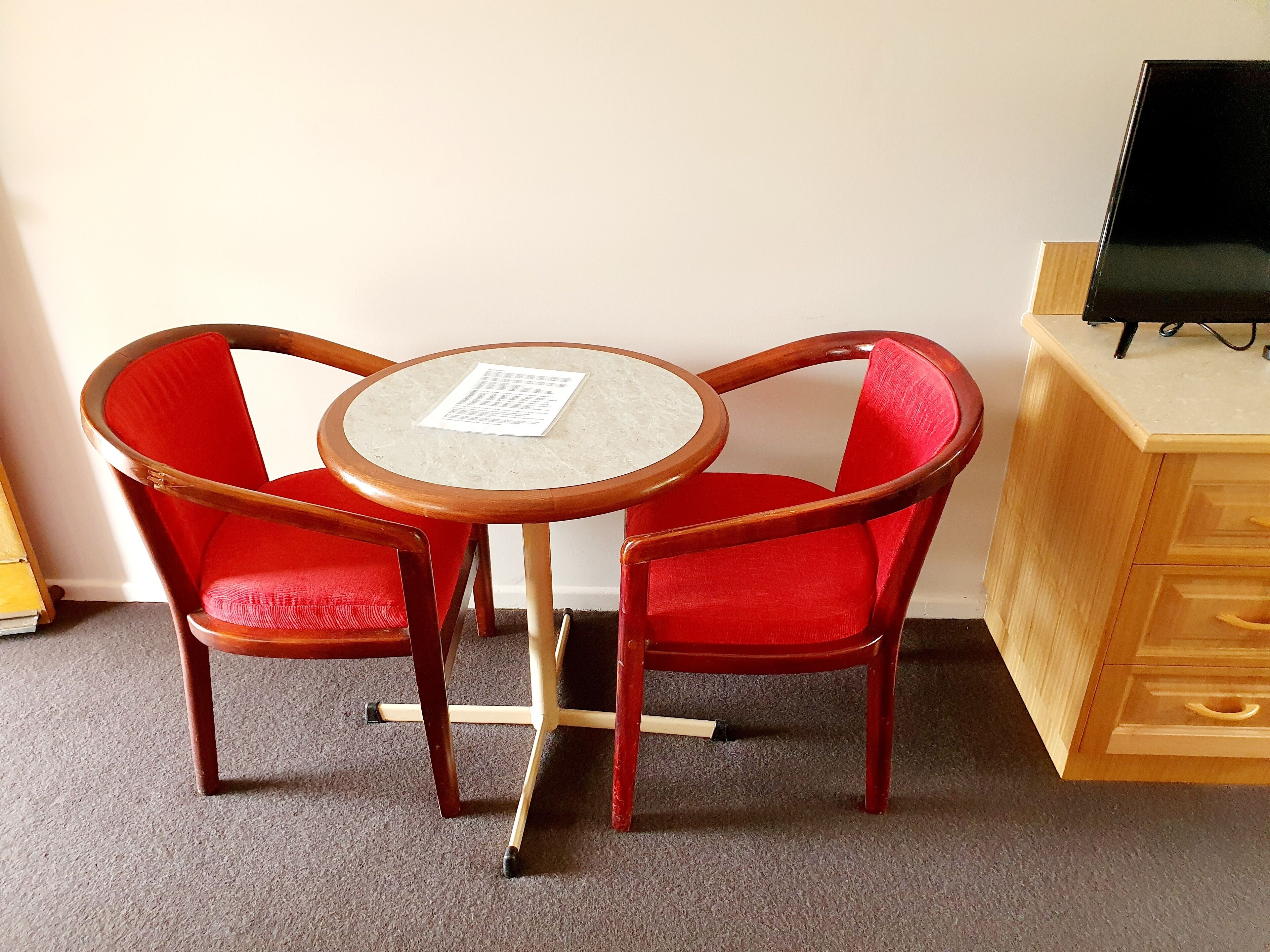 A small room with a round wooden table and two red upholstered chairs. A piece of paper is on the table.