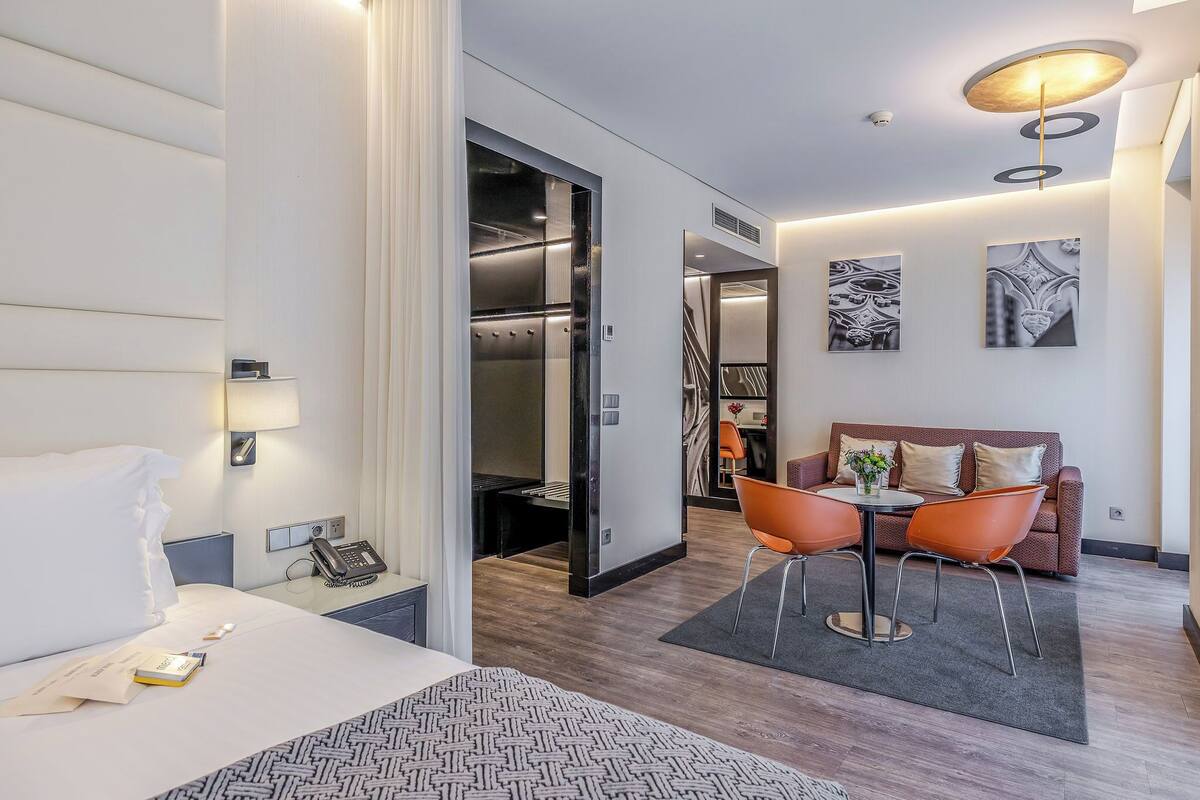 A hotel room with a bed, a desk with a phone, a seating area with a table and chairs, a sofa, and artwork on the walls.