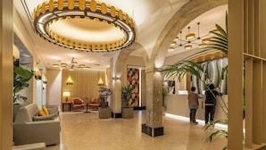 Um hall com um grande candelabro circular, área de estar, balcão de recepção e plantas em vasos.