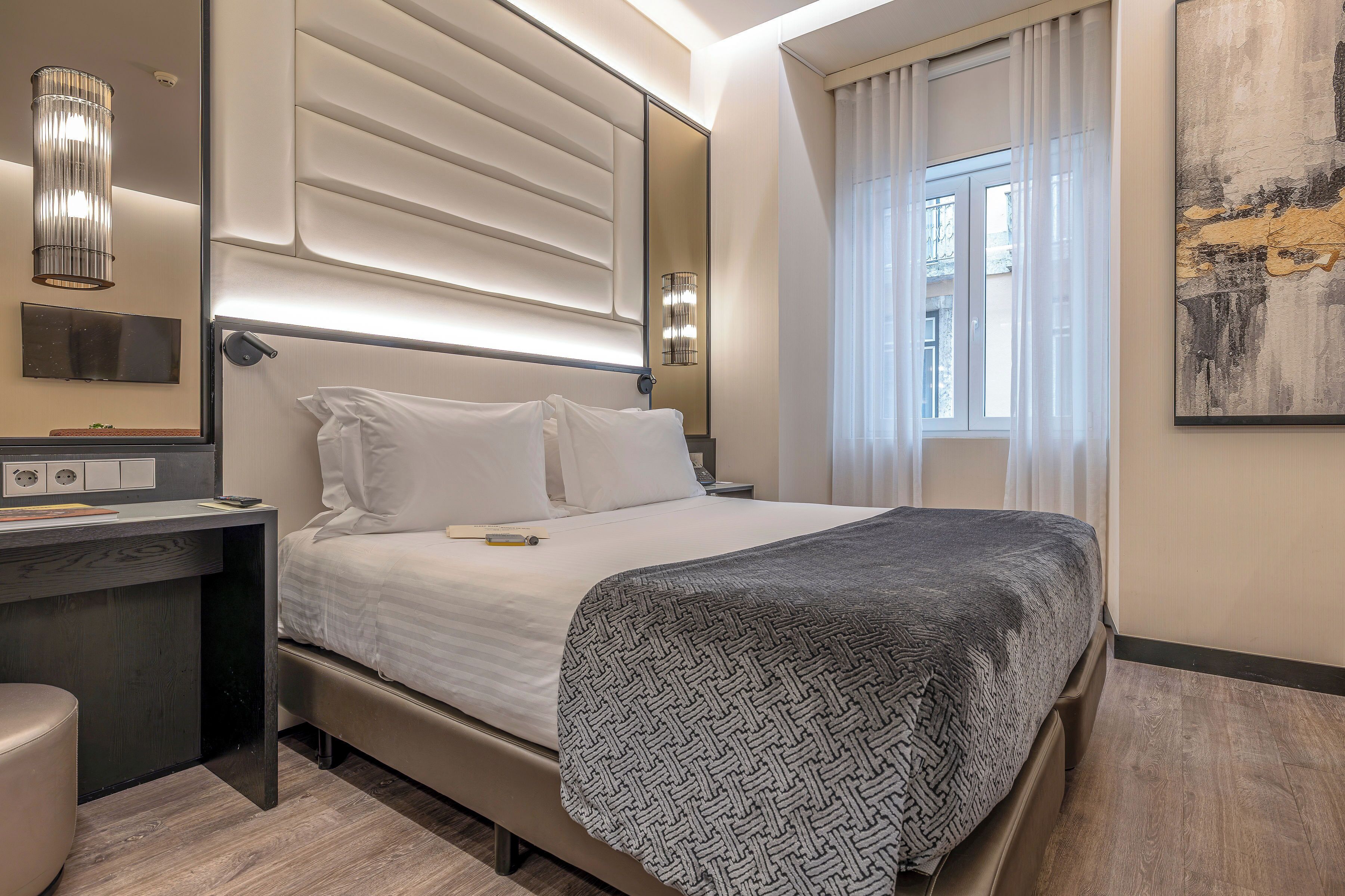 Een moderne hotelkamer met een bed, nachtkastje, lamp en een groot raam met gordijnen.