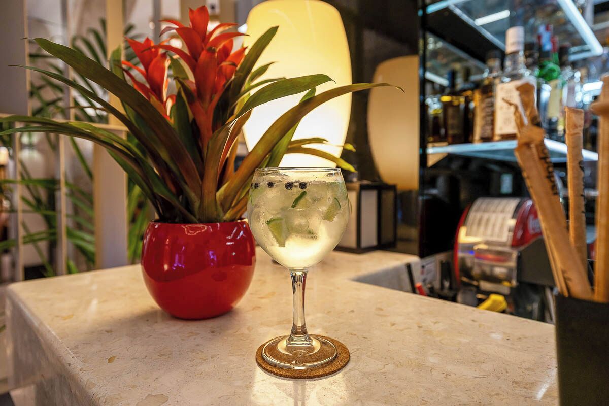 A bar area with a glass of a clear beverage, a red vase with a plant, and shelves with bottles in the background.
