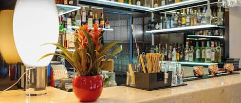 Uma área de bar com prateleiras de garrafas de bebidas, um vaso vermelho com planta e um porta-copos com canudos.