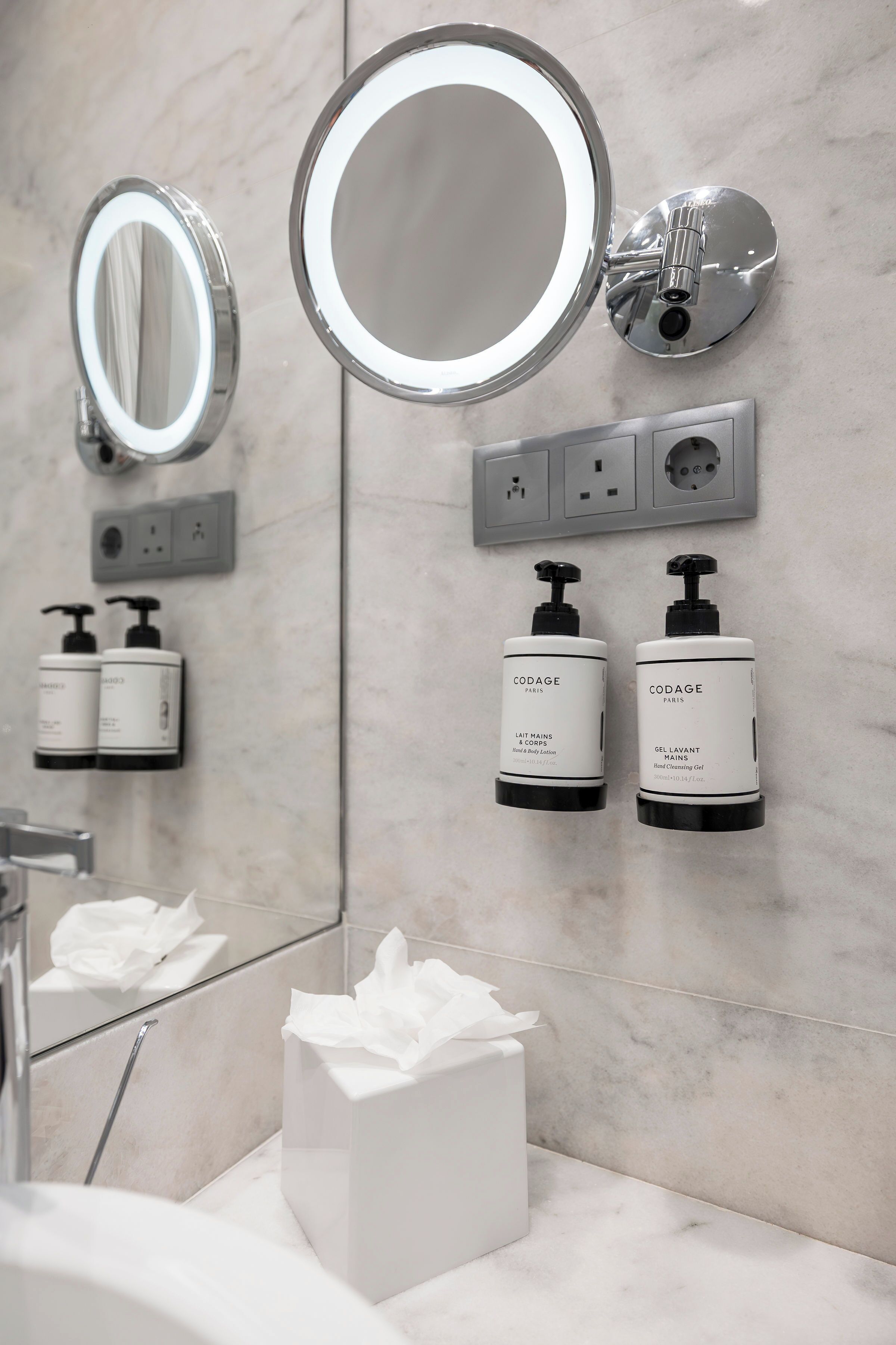 A bathroom with a mirror, two soap dispensers, a tissue box, and a wall-mounted light fixture.