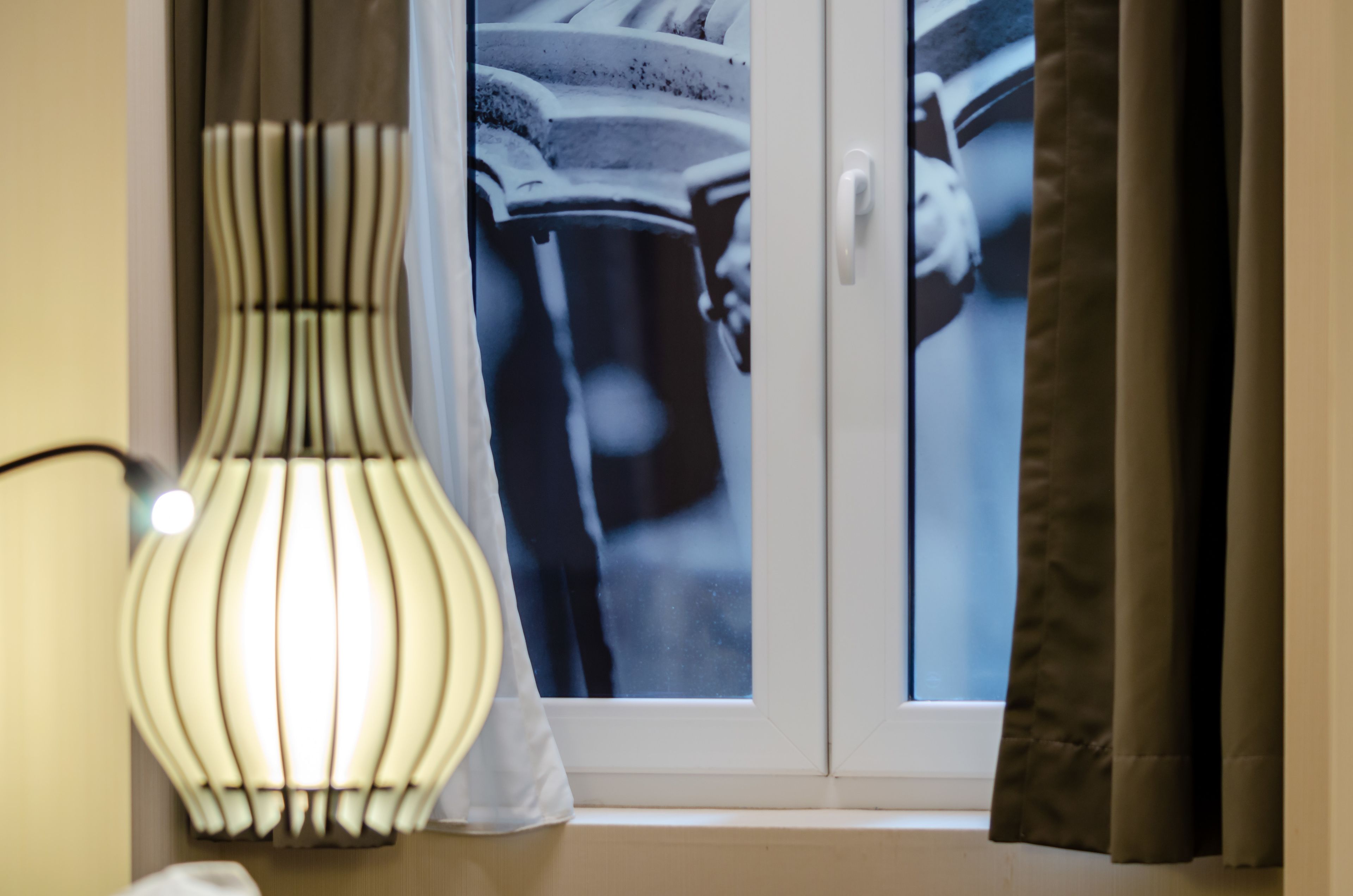 A window with a view of a person in a white coat, curtains, and a lit lamp.