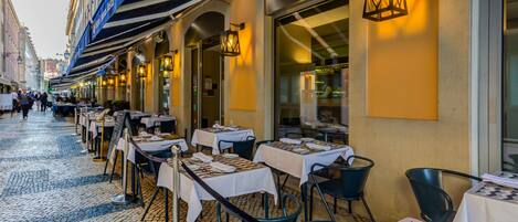 Un área de comedor al aire libre con mesas dispuestas para los comensales, que presenta un suelo adoquinado y una fila de mesas con sillas.