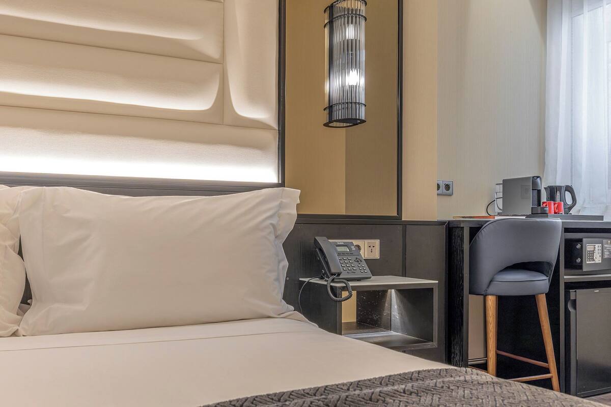 A hotel room with a bed, bedside phone, desk with a chair, and a coffee maker.