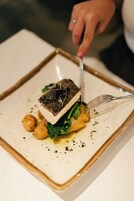 A square plate with a piece of fish on top, surrounded by greens and small round items, with a fork and knife on the side.