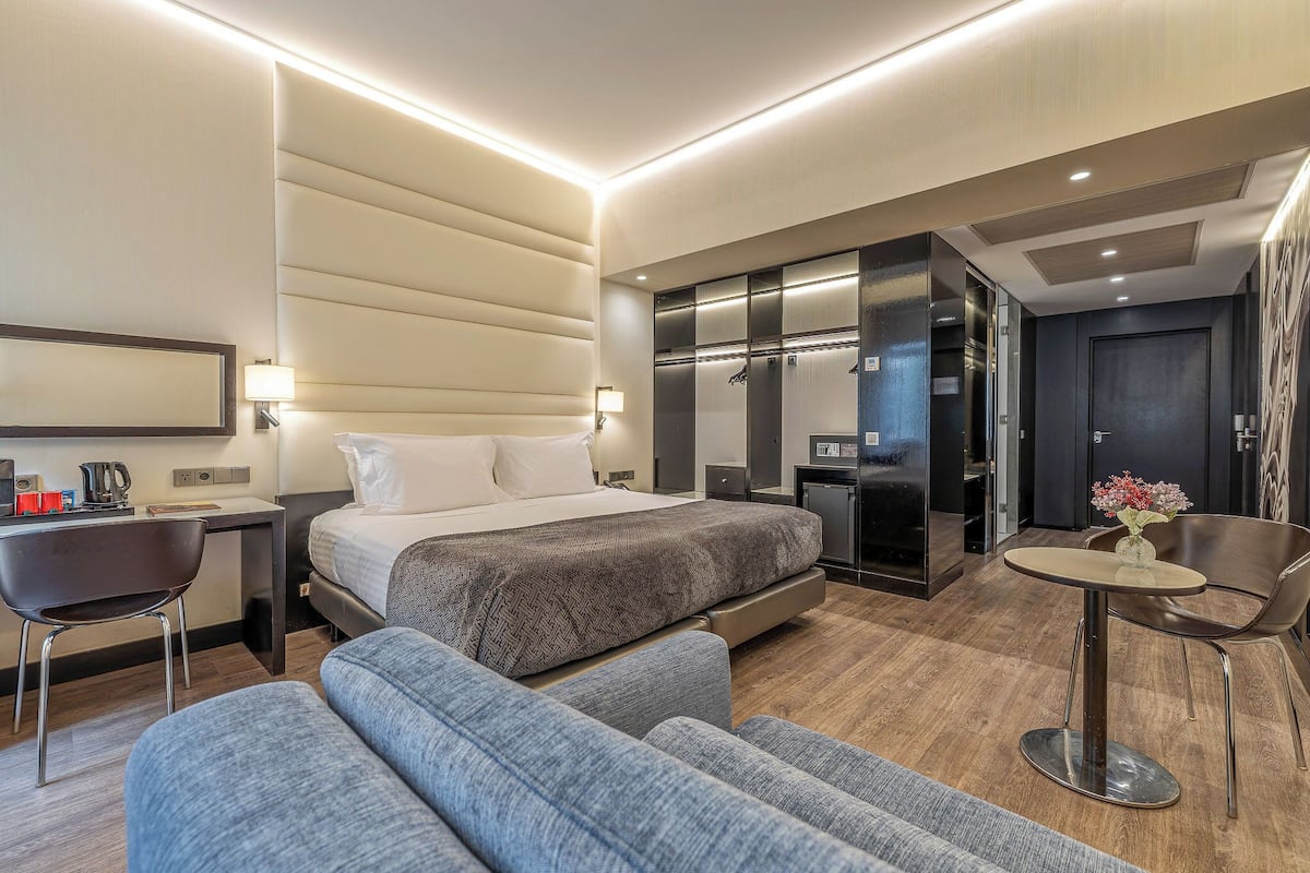 A modern hotel room with a bed, a desk, a chair, a sofa, and a small table with a vase of flowers.