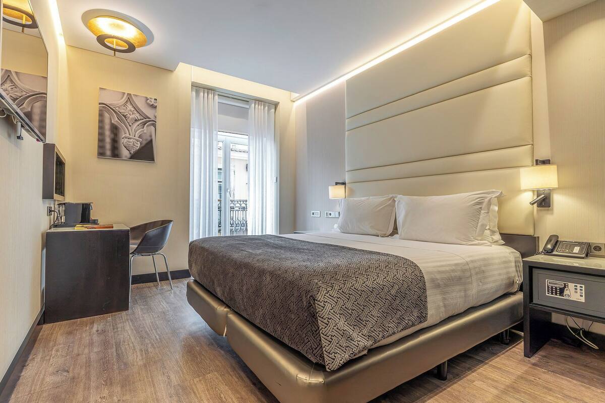 A modern hotel room with a large bed, bedside table, and a desk with a chair.