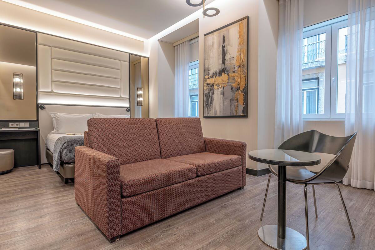 A modern hotel room with a sofa, a small round table, a bed, a desk, and a large abstract painting.