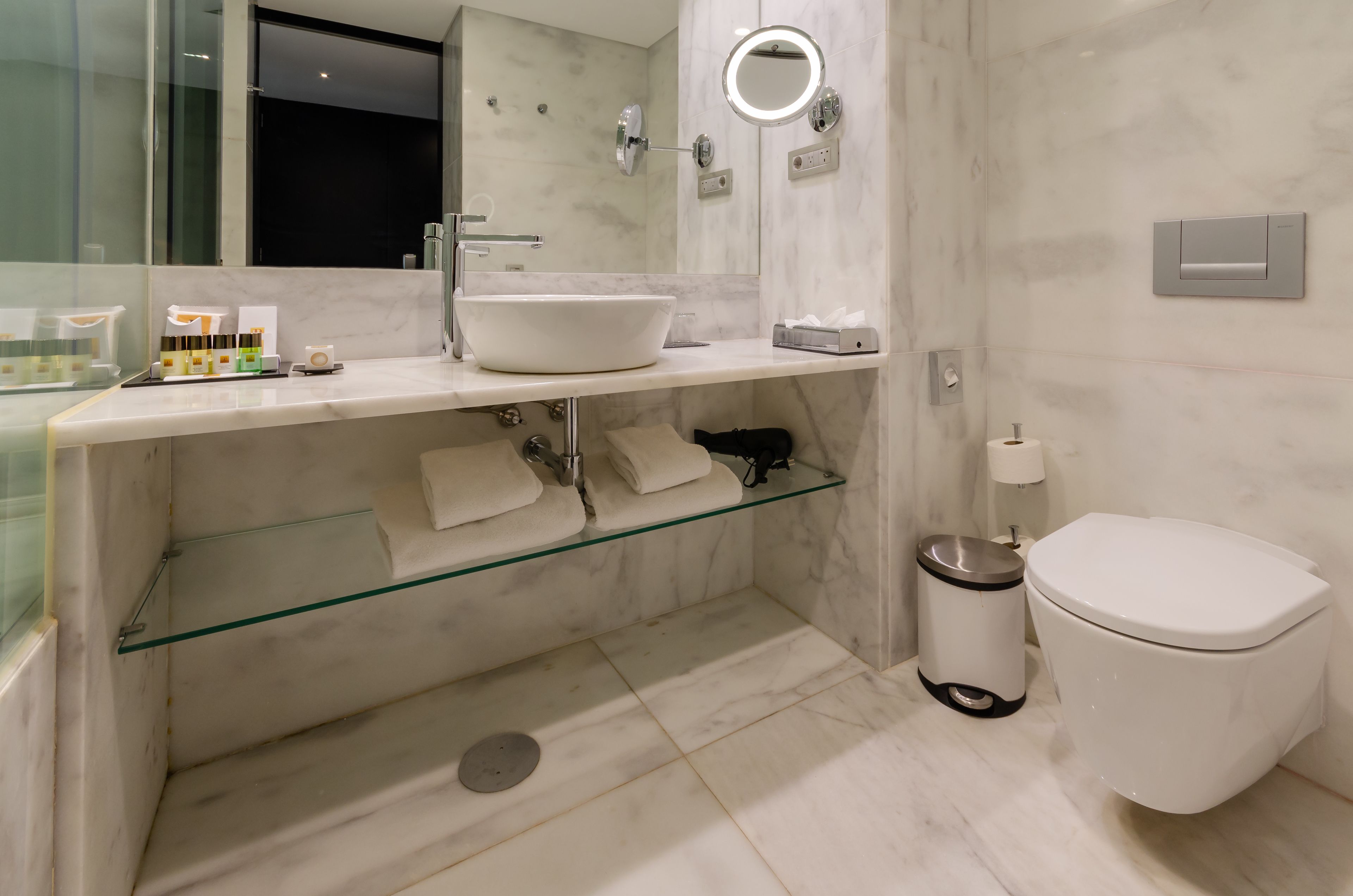 Un baño moderno con lavamanos blanco, estante de vidrio y un inodoro.