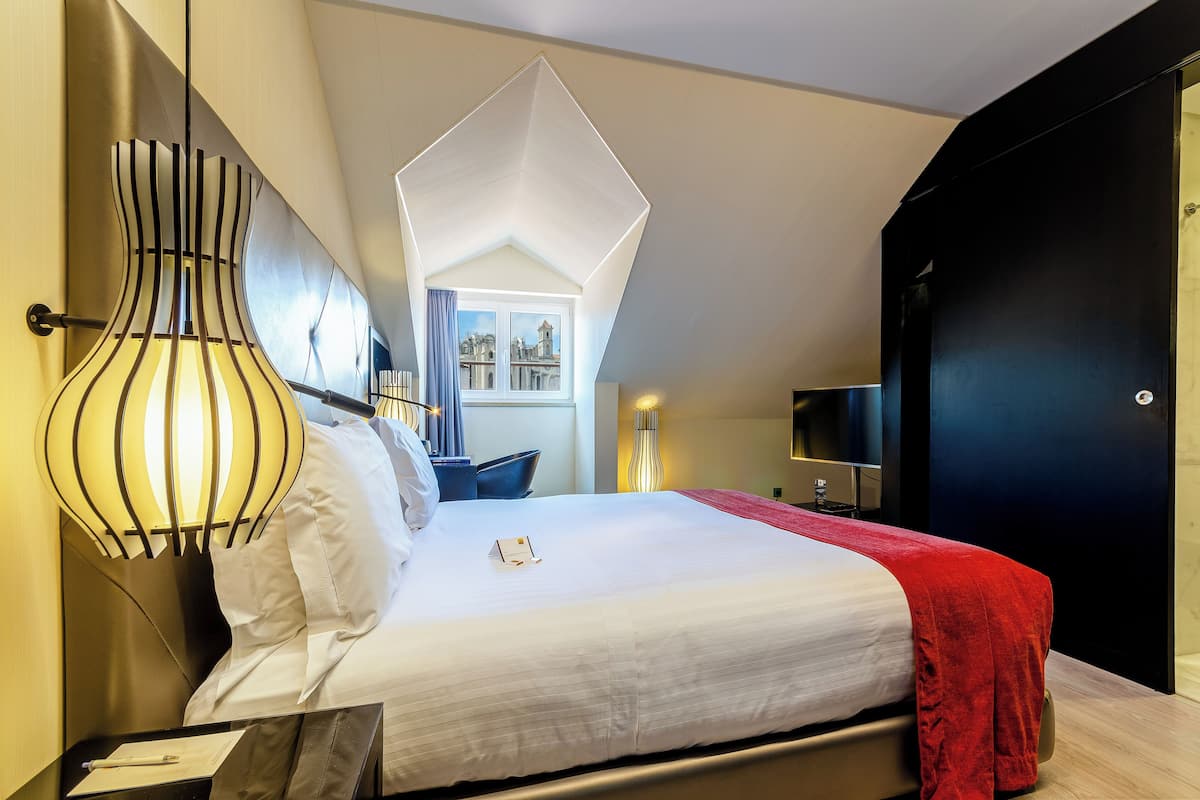 A modern hotel room with a neatly made bed, a unique geometric ceiling light, and a view of the city through a window.